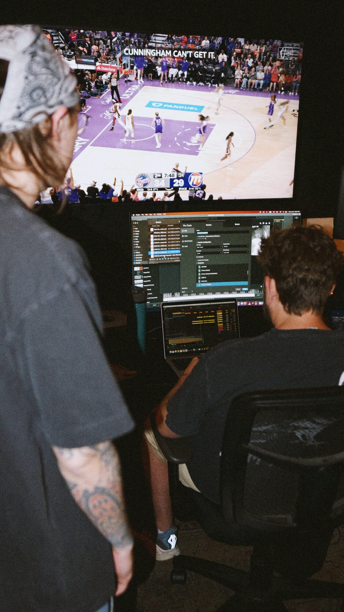 Two people working on computer screens with a basketball game visible on a large monitor in the background.