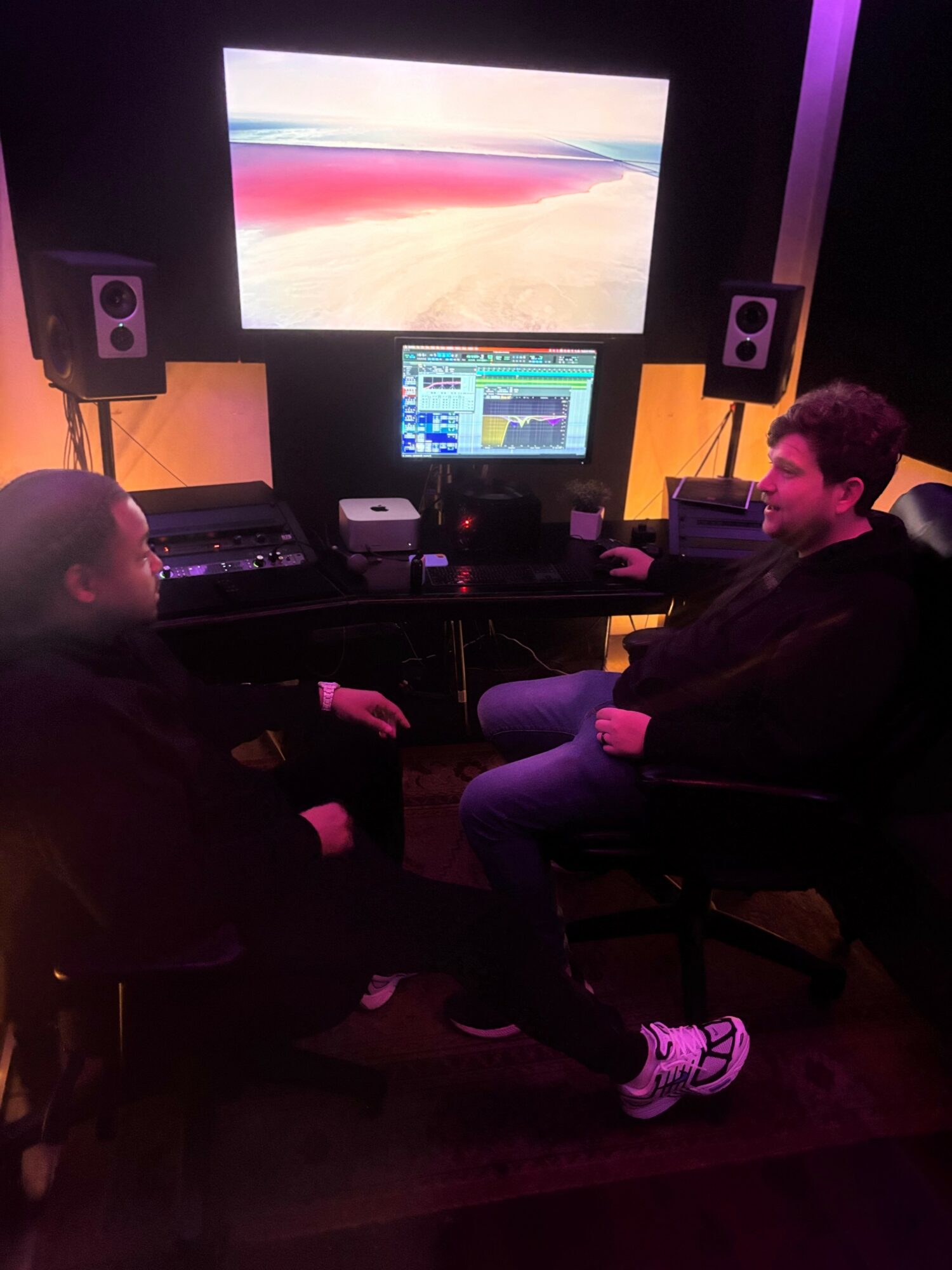 Two people sitting in a music studio with a large screen, speakers, and a computer setup, facing each other and smiling.