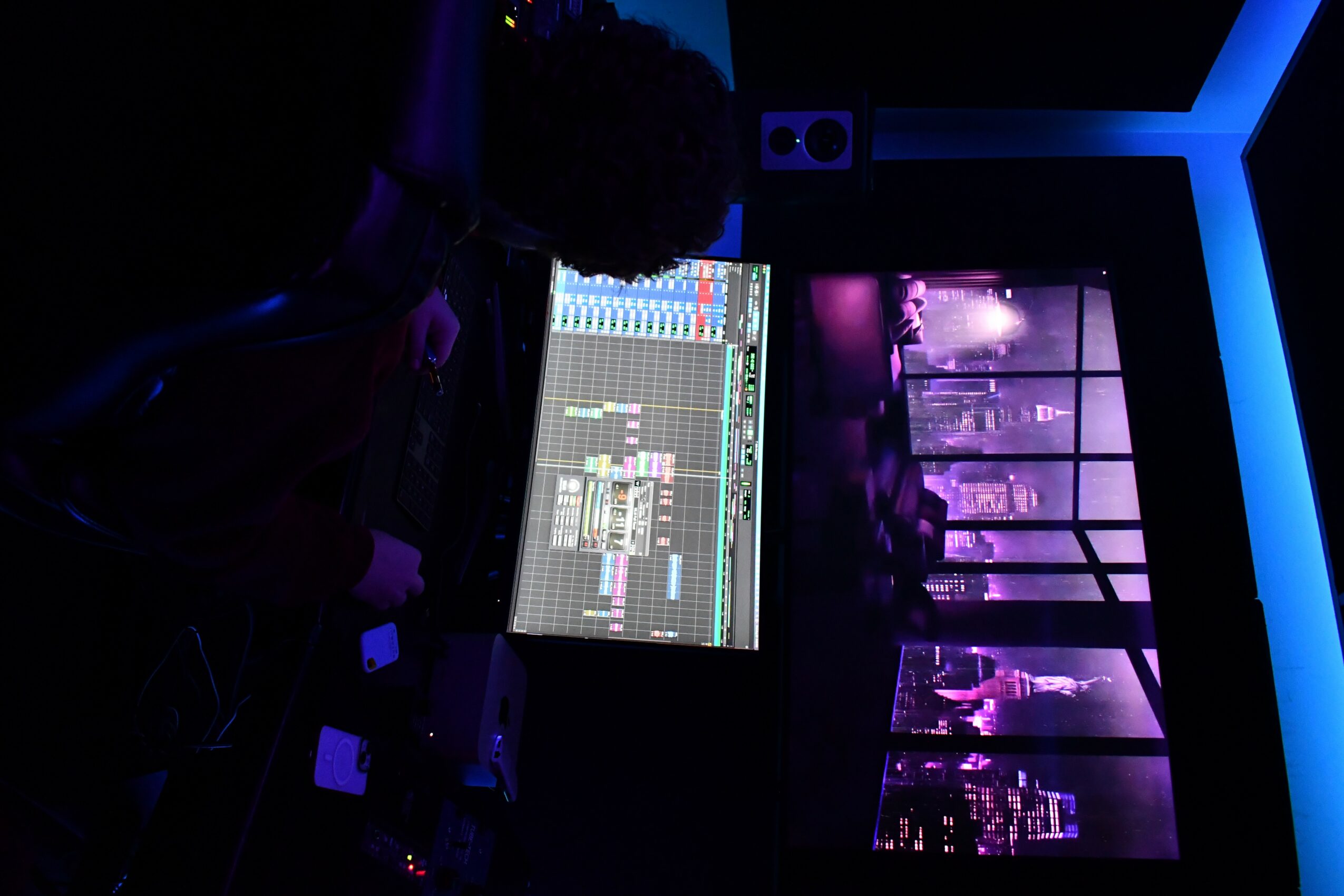 A person working on a music production computer in a dimly lit room with purple and blue lighting, city skyline view through windows.