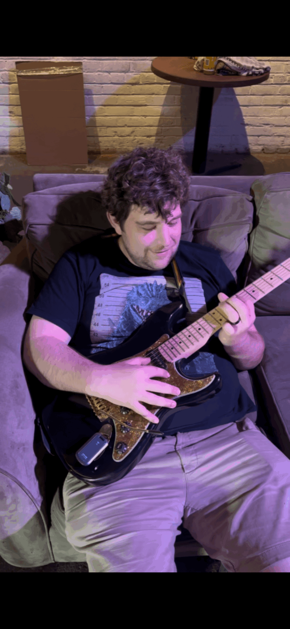 Young person with curly hair playing an electric guitar on a couch, with a table and a cardboard box in background.