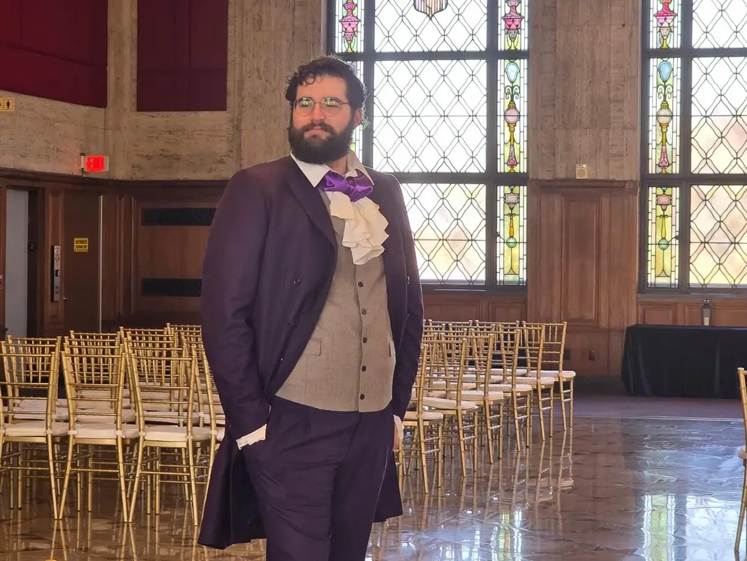 Man with beard in formal attire standing in a decorated hall with stained glass windows and chairs.