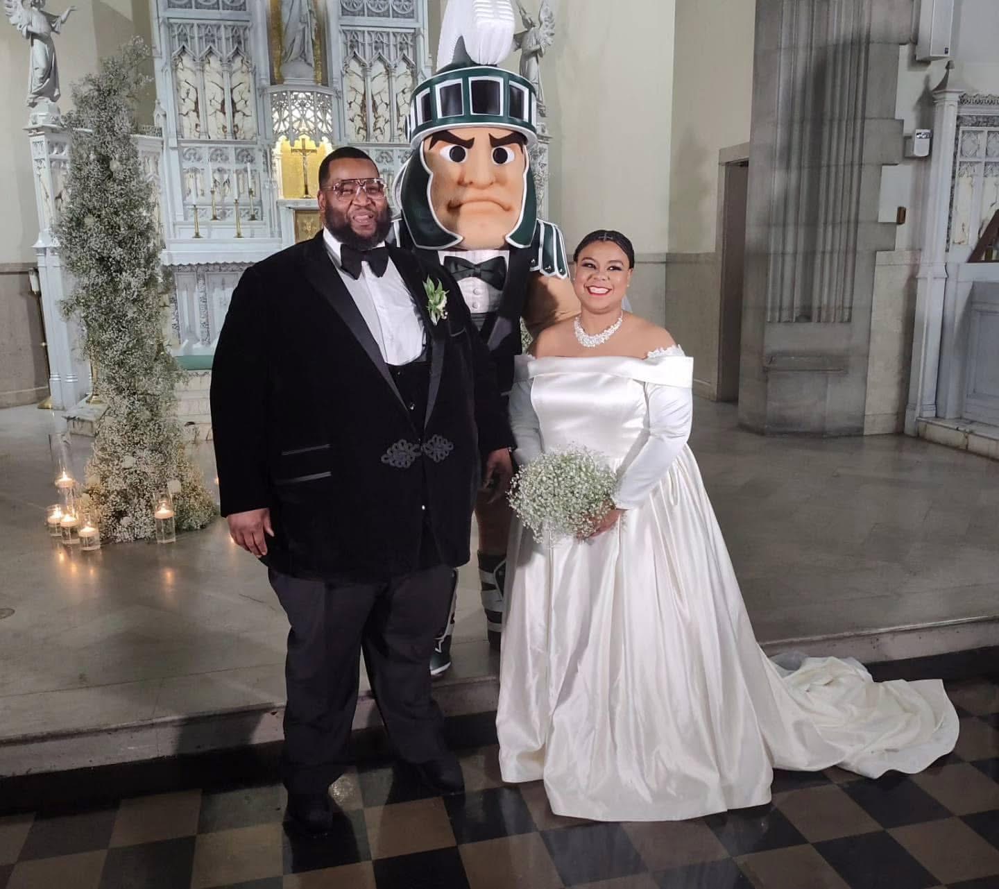 A bride in a white wedding dress holding a bouquet, a man in a tuxedo, and a person in a knight costume pose indoors.