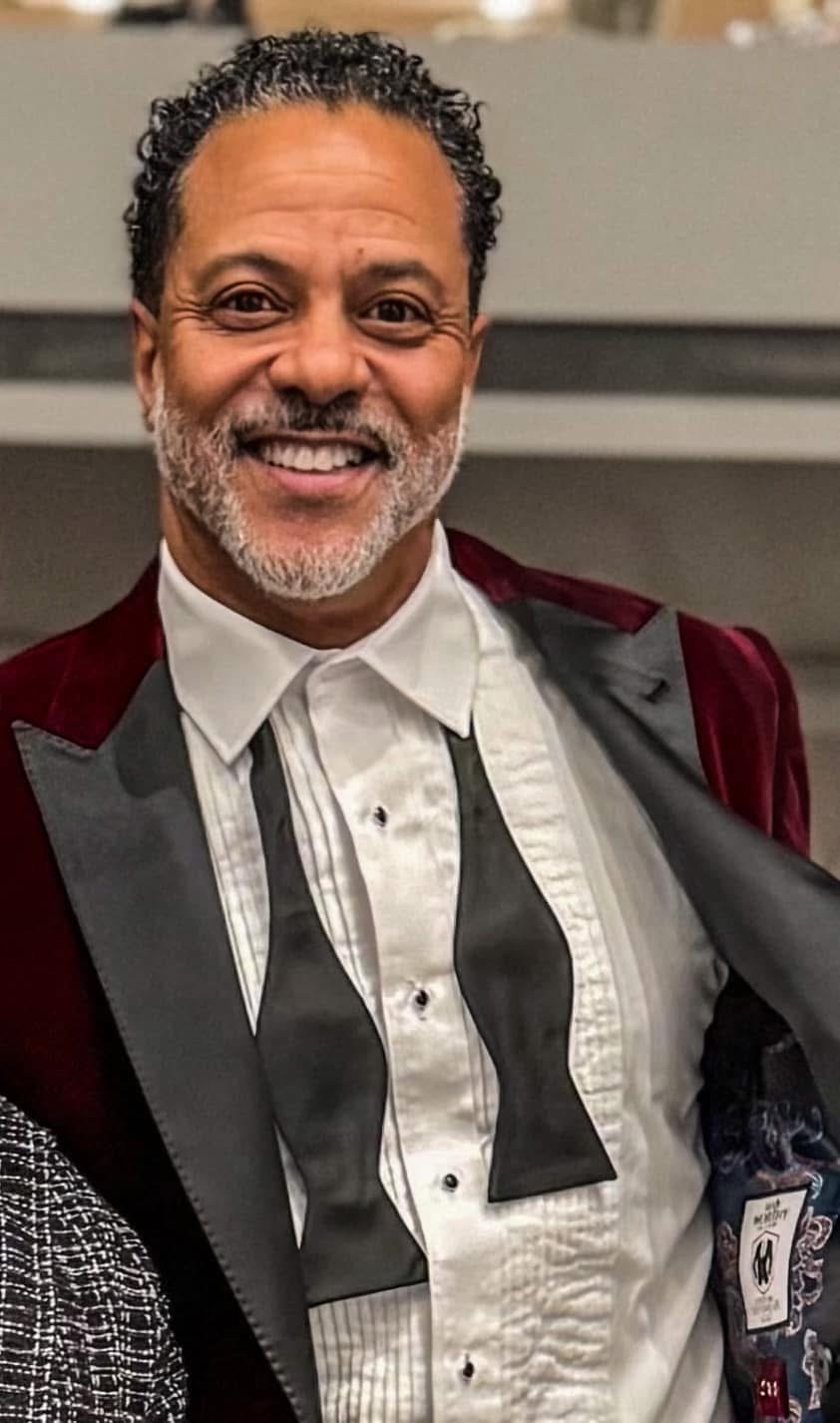 Smiling man with curly hair and beard wearing a tuxedo with a bow tie, standing indoors.