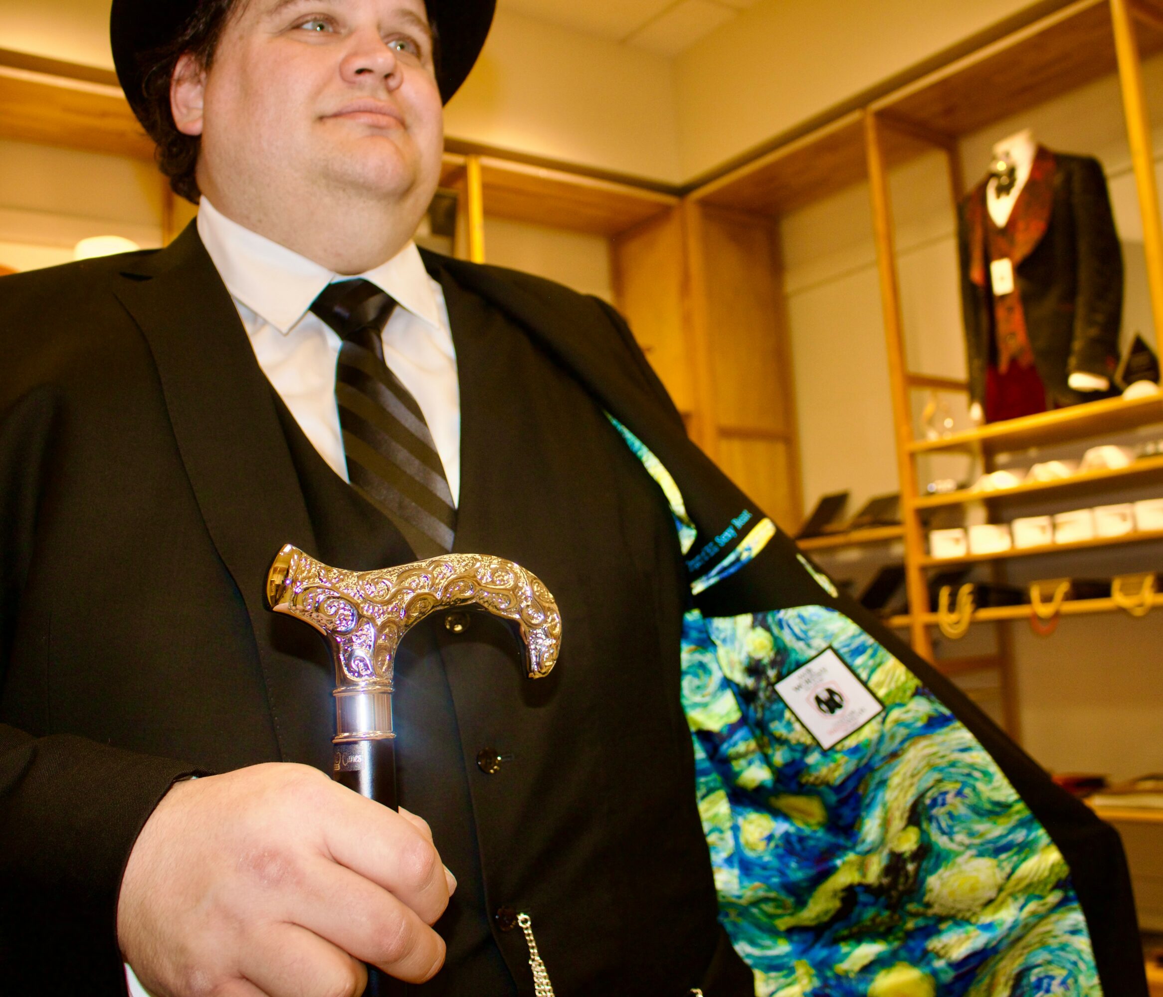 Person in formal attire holding a cane, with a colorful jacket and a display of suits in the background.