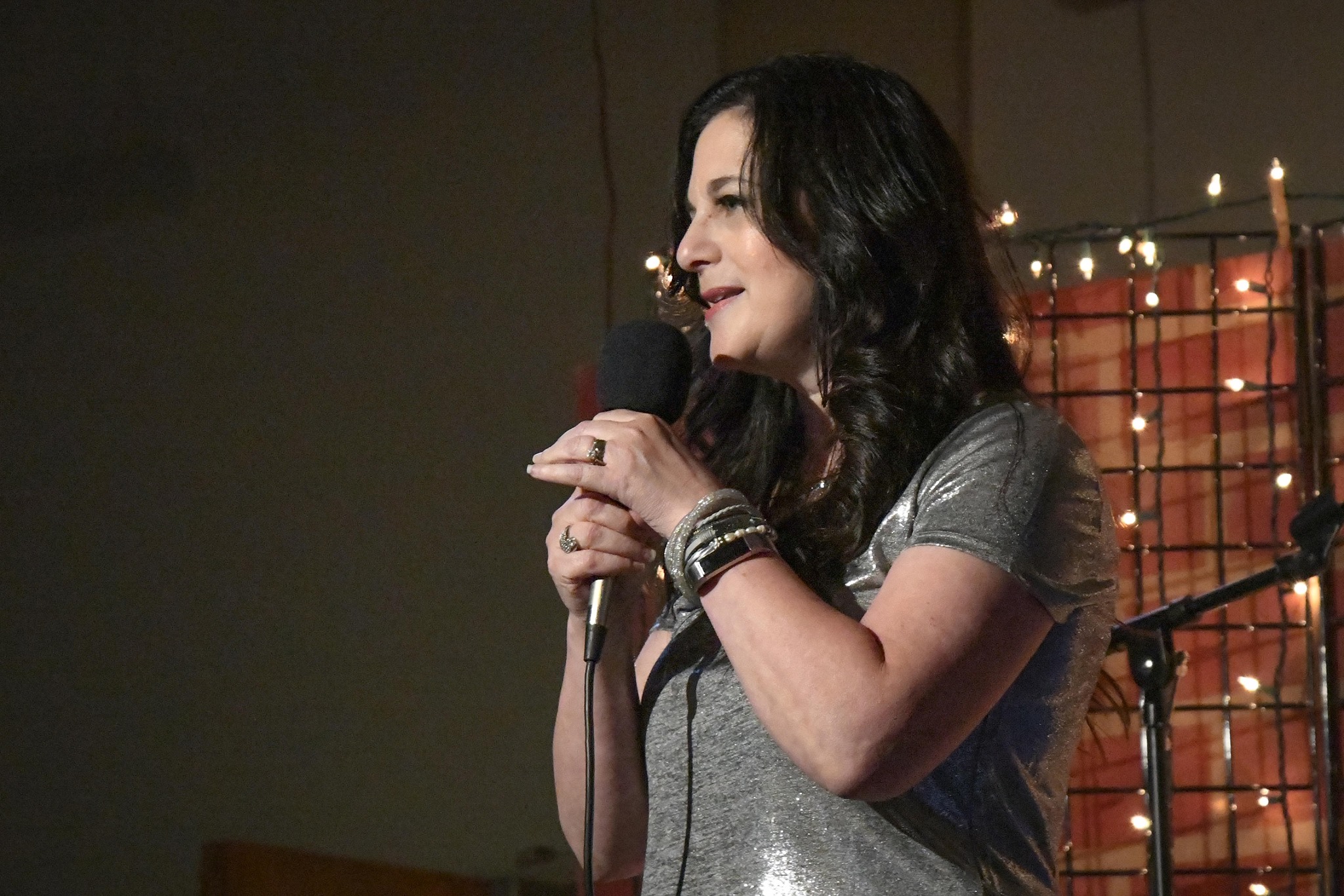 Woman with dark hair holding a microphone, standing on stage with string lights in background.