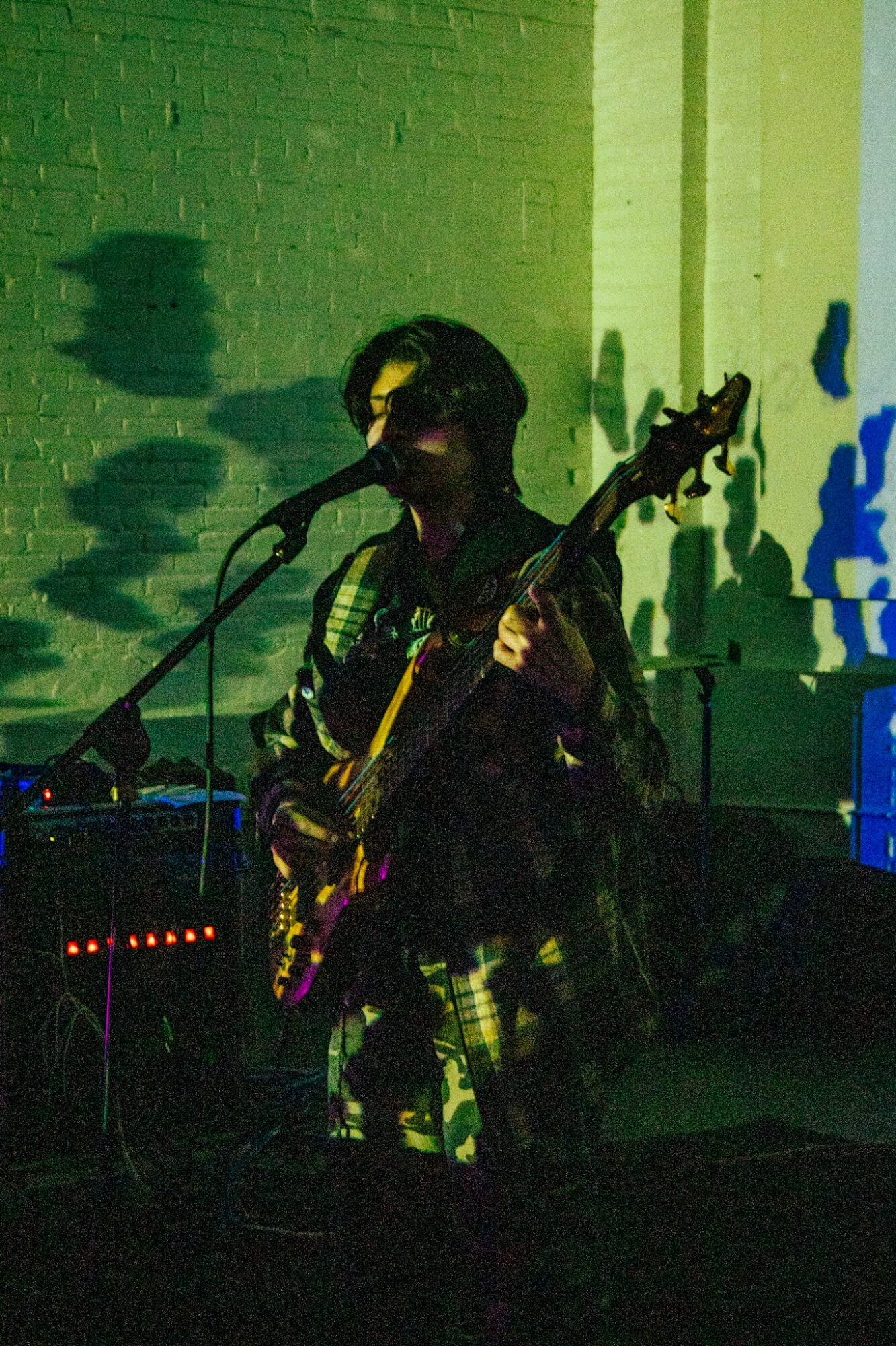 Person playing guitar on stage with green and blue lighting, shadow cast on brick wall behind.