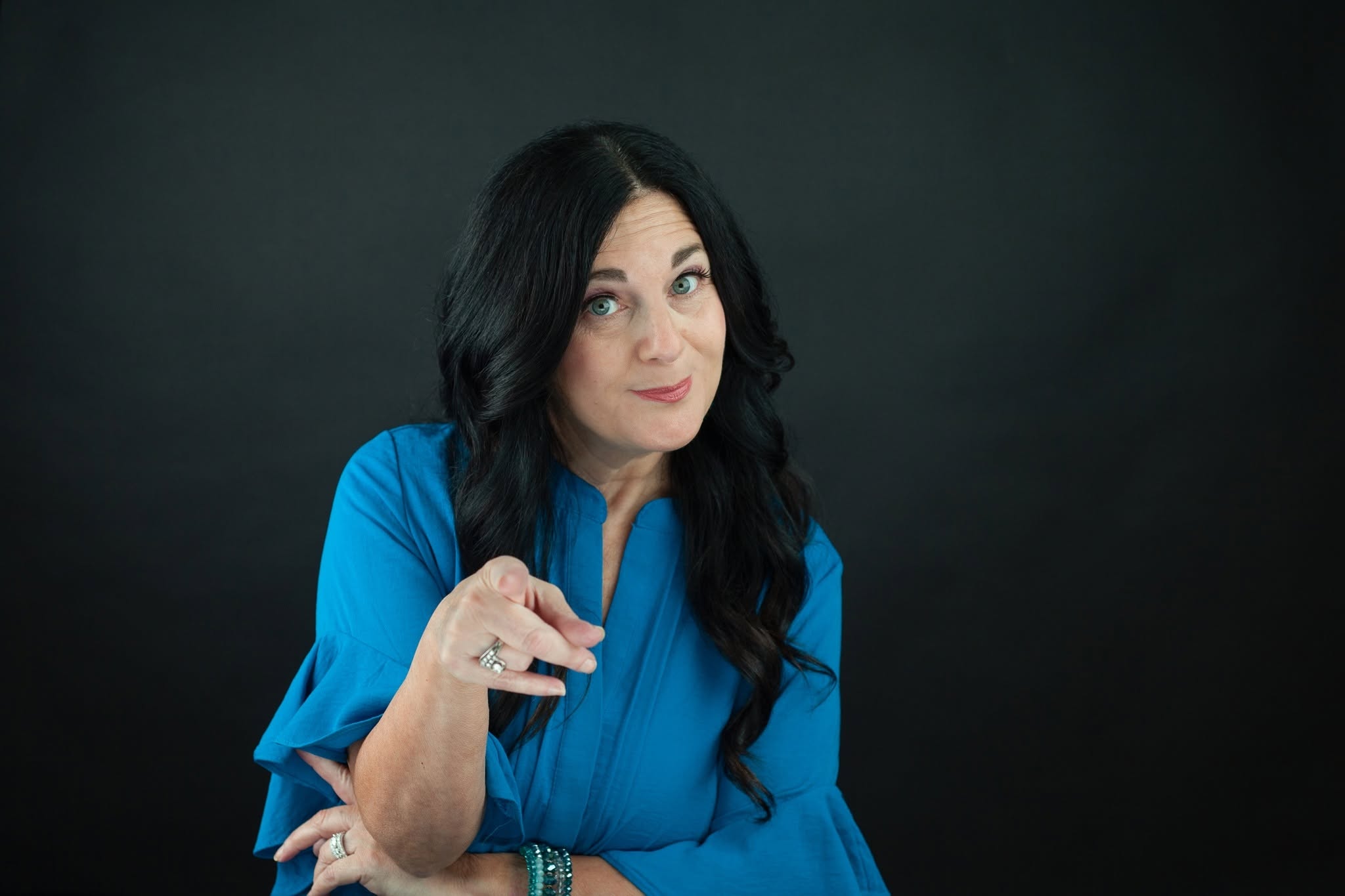 Woman with long dark hair wearing a blue top, pointing with her right hand, against a dark background.
