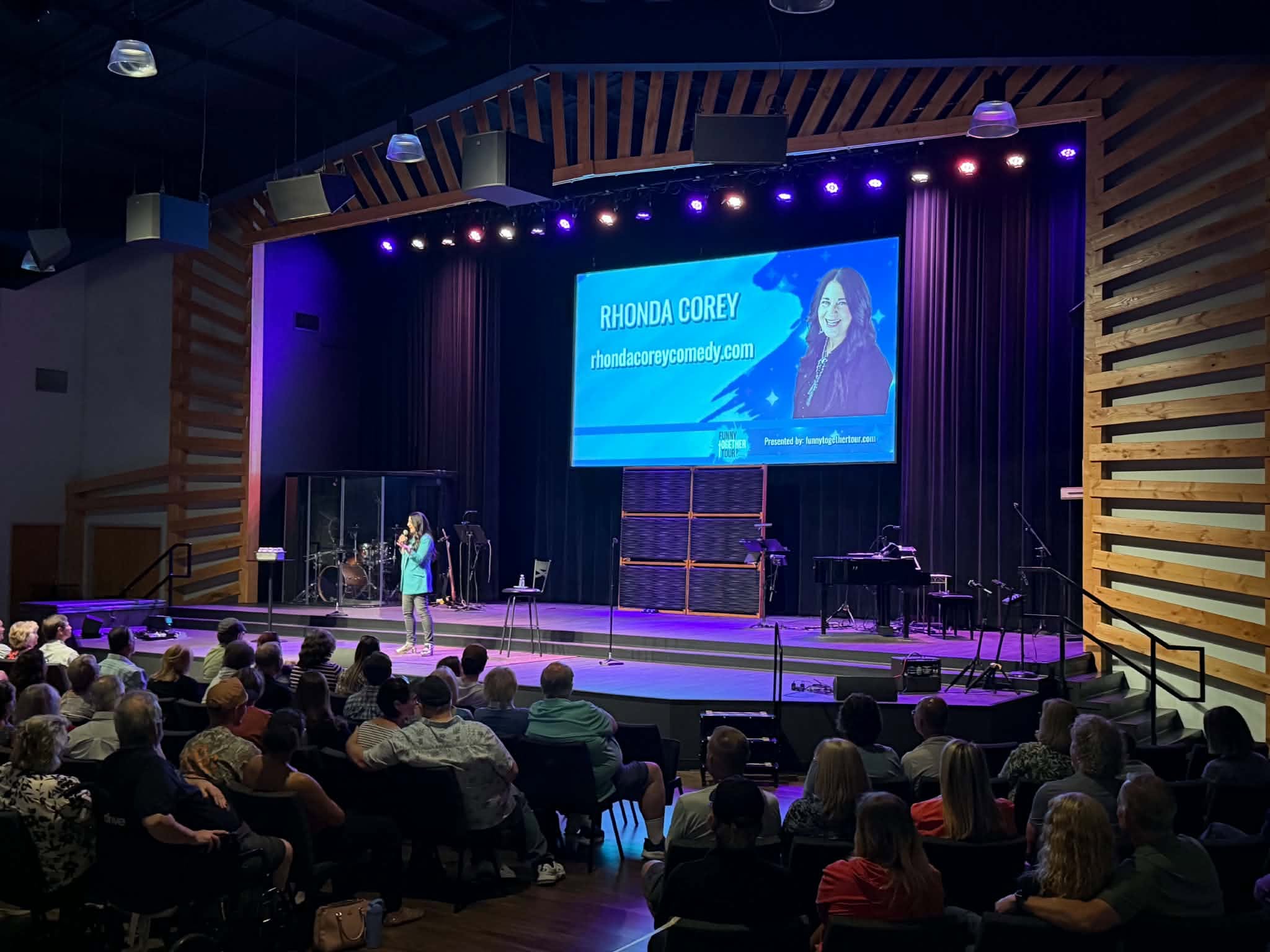 Audience seated in a theater watching a presentation on stage with a large screen displaying a woman and text.