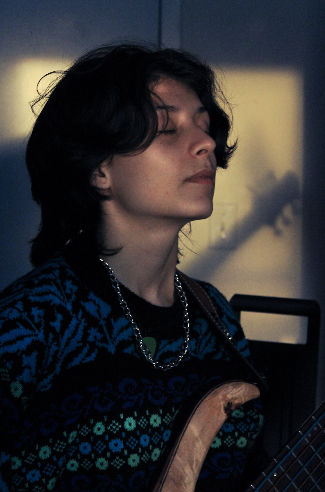 Side profile of a person with dark hair, wearing a patterned top and necklace, in a dimly lit room.
