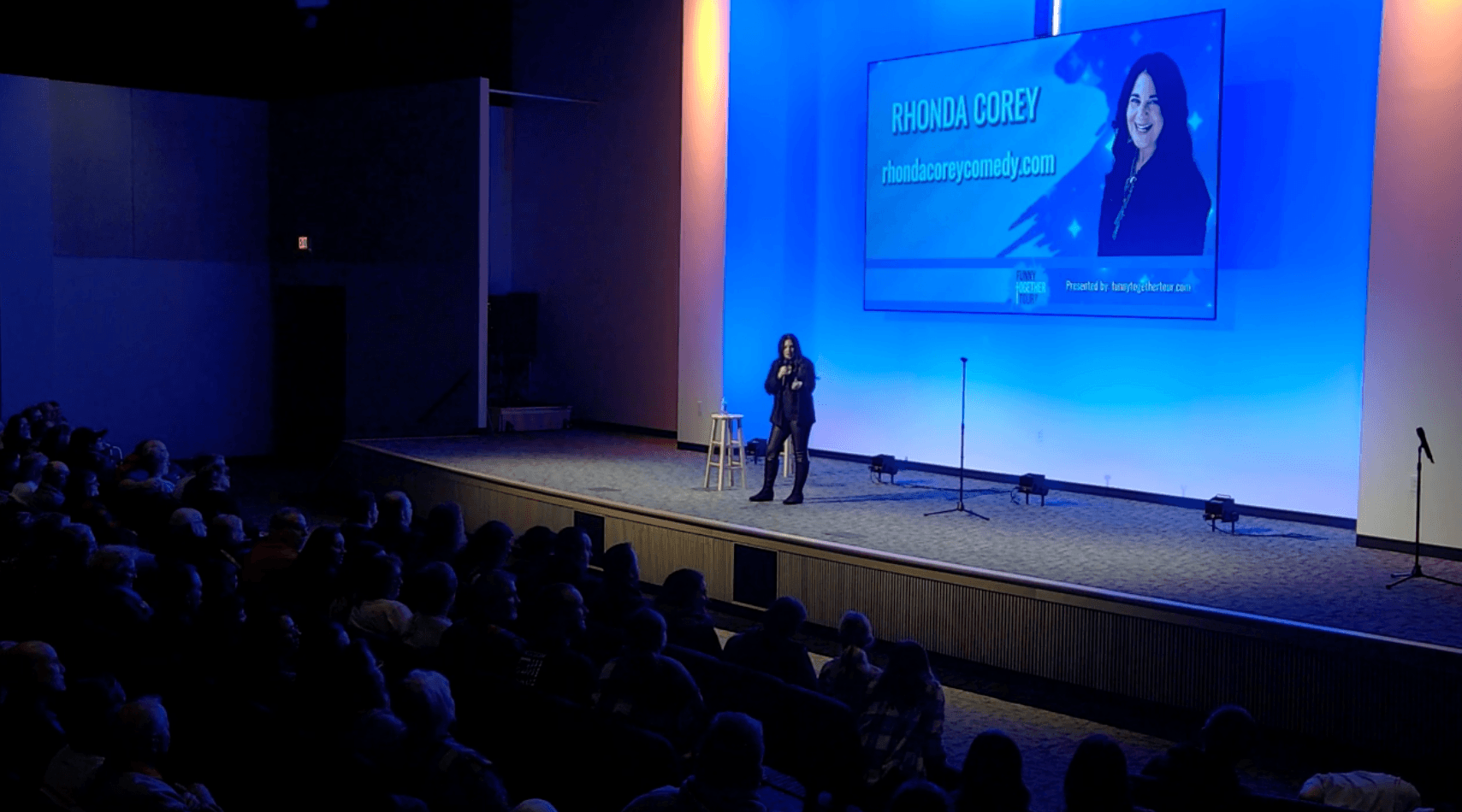 Person speaking on stage in front of a large screen with an audience watching, stage lit with blue lighting.
