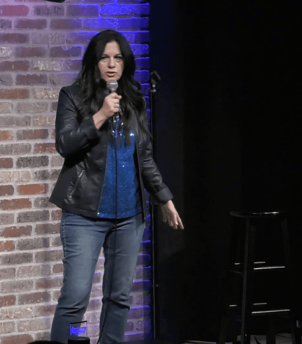 Woman with long dark hair holding a microphone on stage, brick wall background, wearing a black leather jacket and blue top.