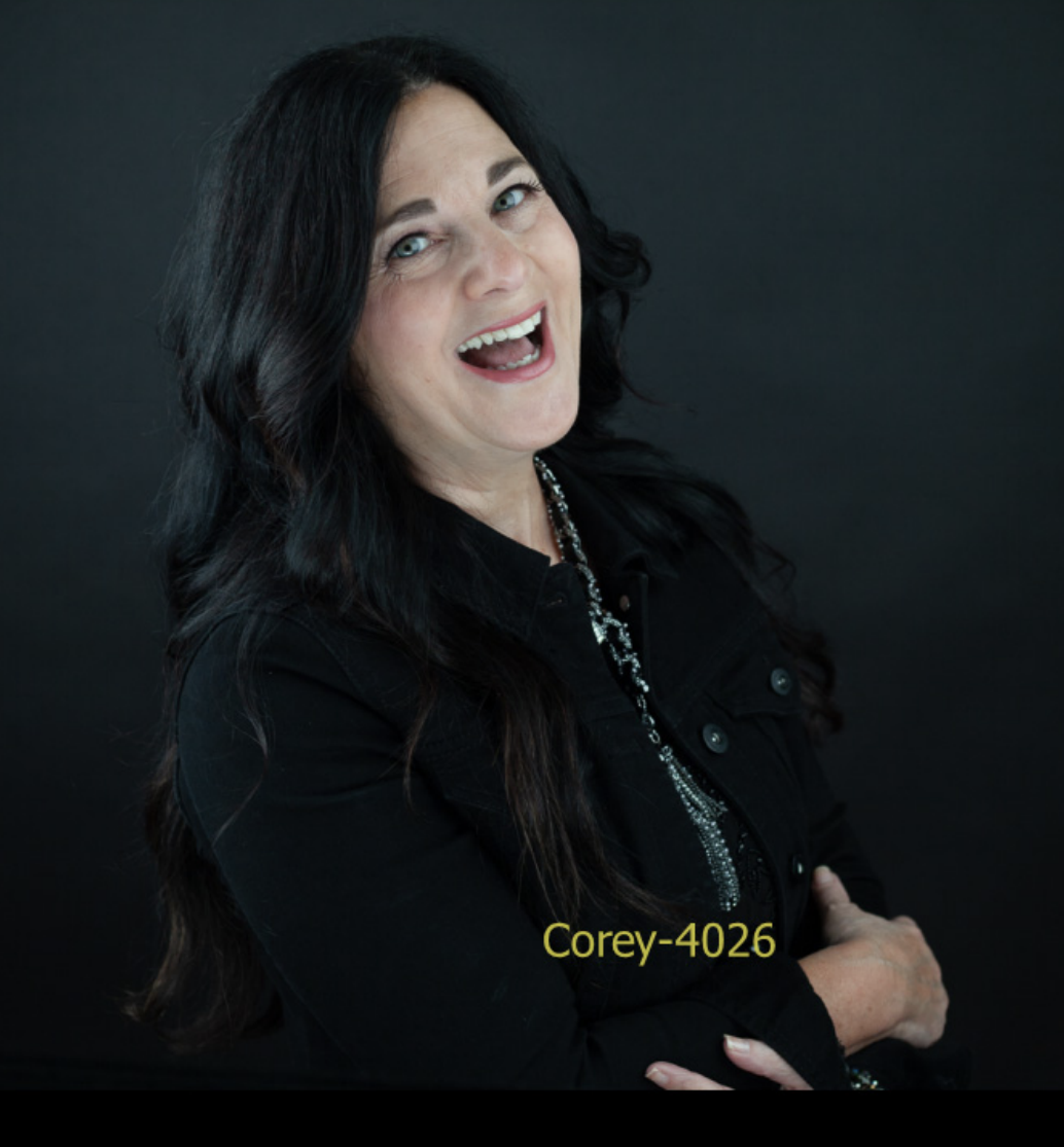 Woman with long dark hair smiling, wearing a black jacket and necklace, against a dark background.