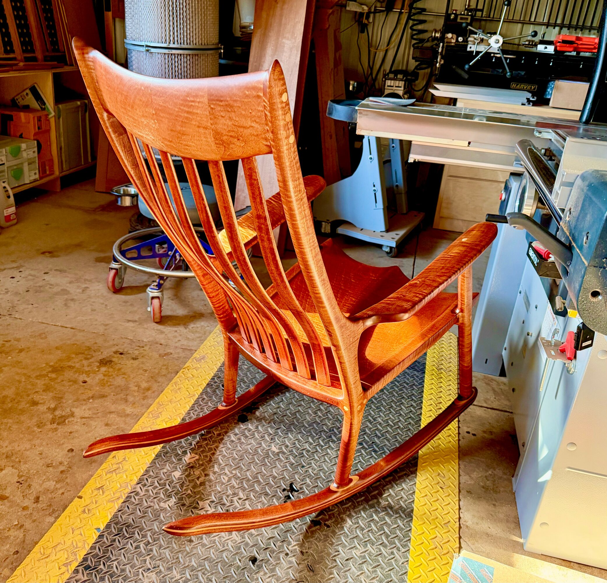 Wooden rocking chair with a high back and slatted design, placed on a textured mat in a workshop.