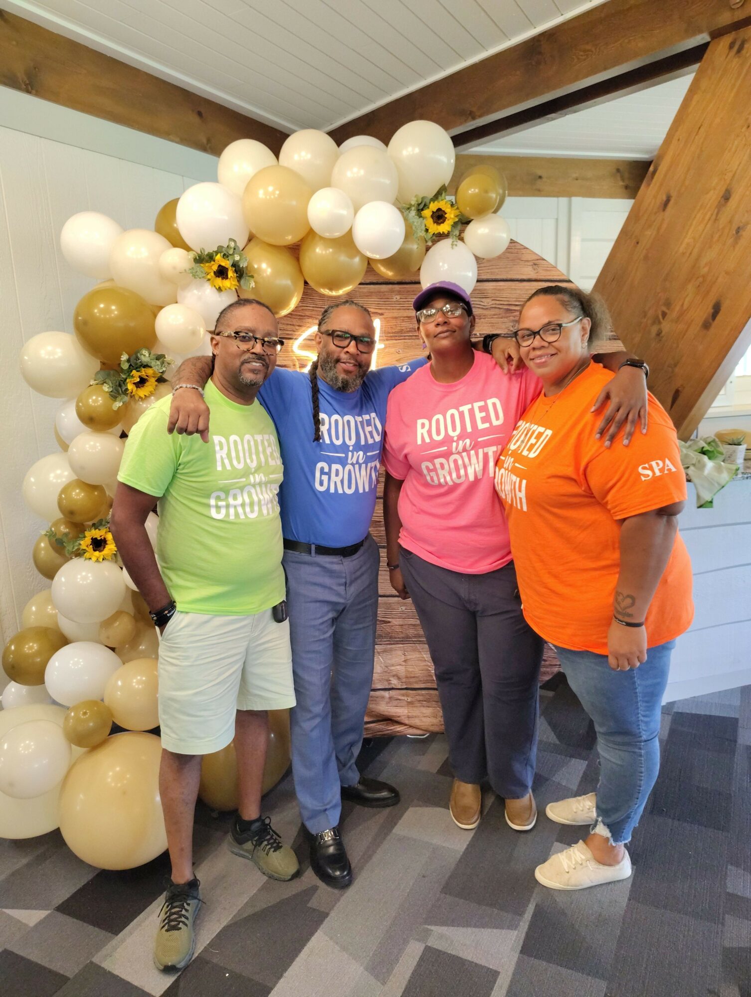 Four people standing indoors near a balloon and sunflower decoration, smiling and posing for a photo.