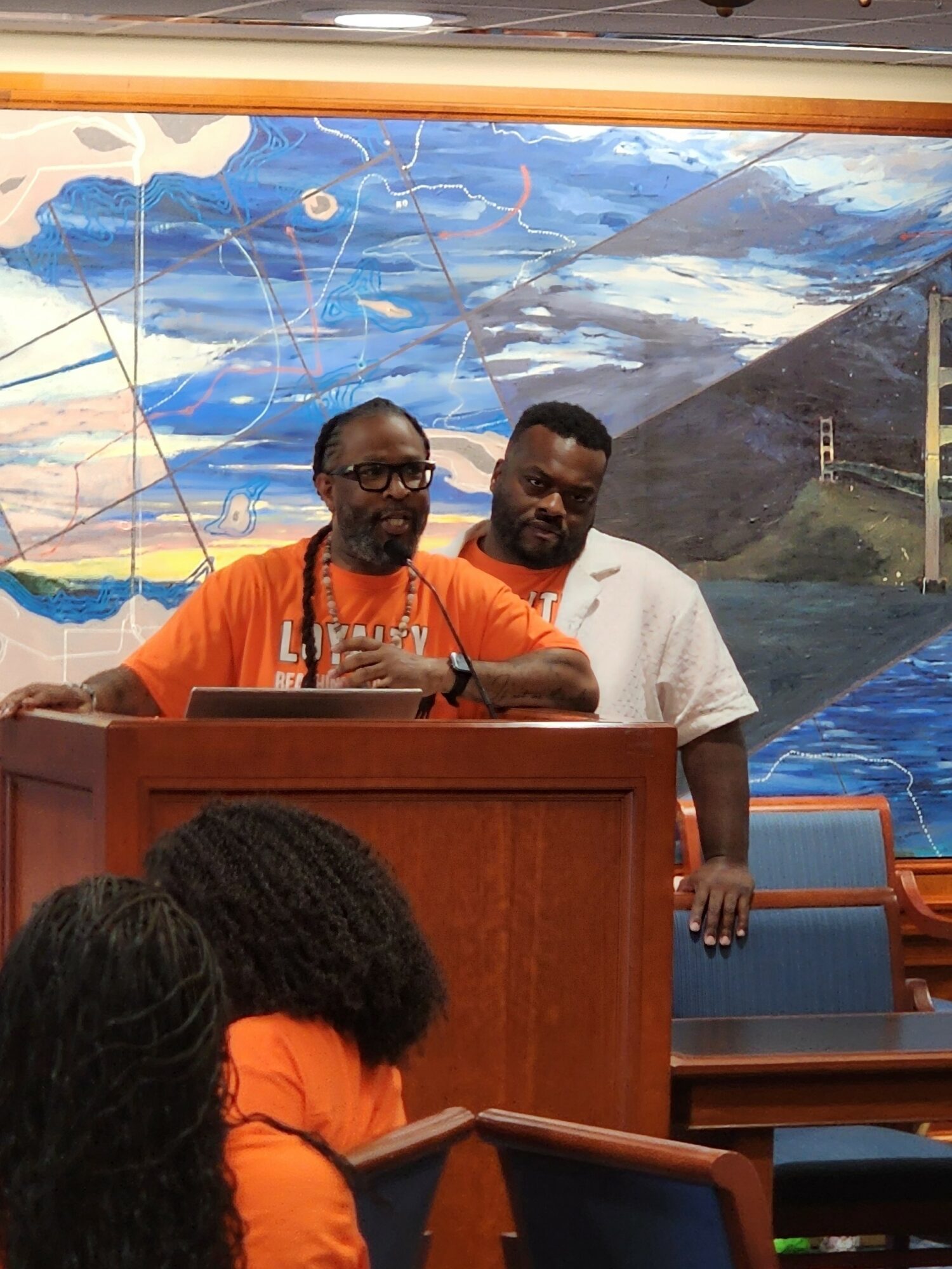 Two men stand behind a podium, one speaking into a microphone, with a colorful mural of sky and landscape in the background.