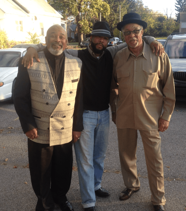 Three men standing outdoors, smiling, with cars and trees in the background.