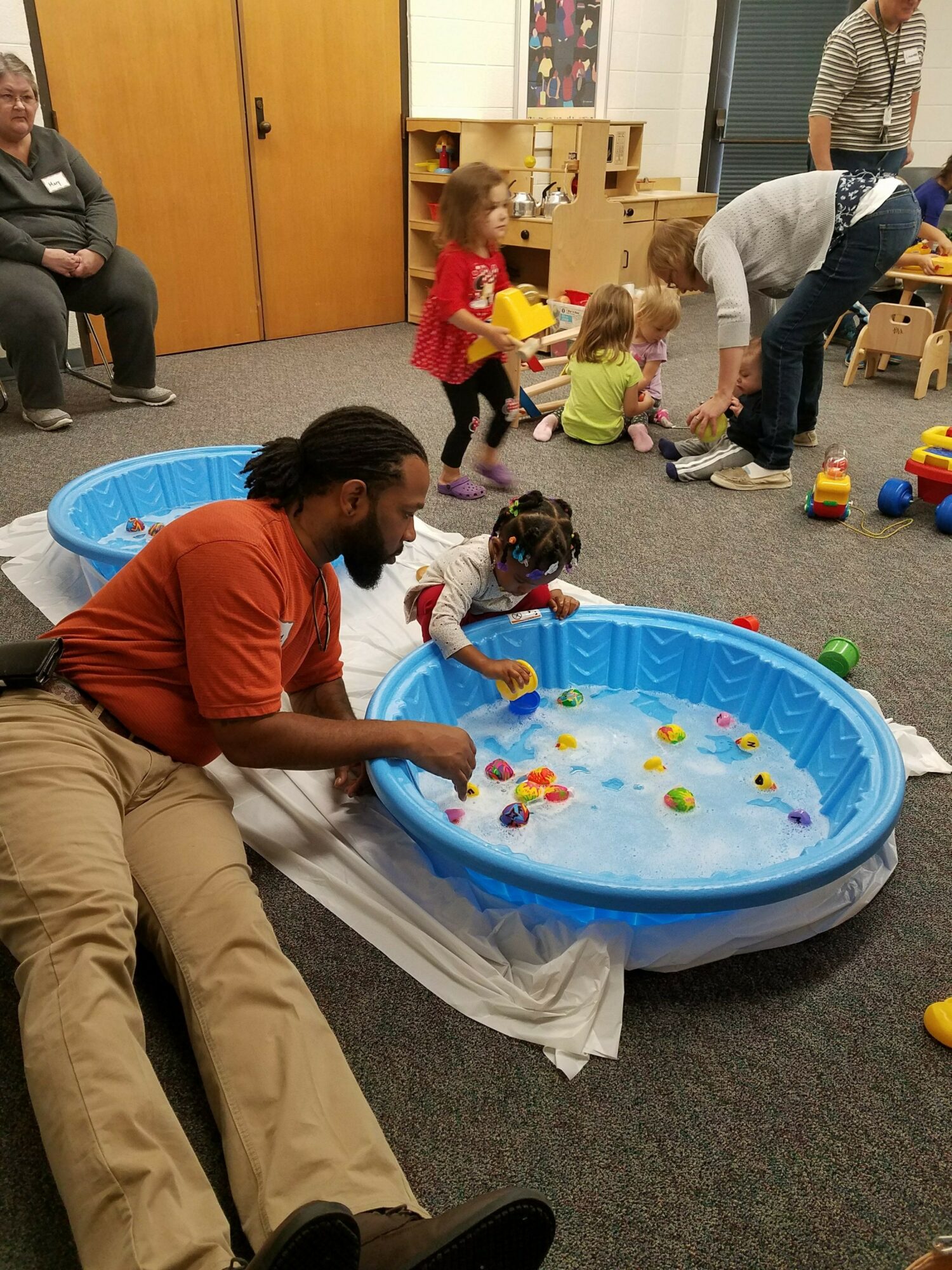 Adult man and children playing with water in a small blue pool indoors, with other children and adults nearby.