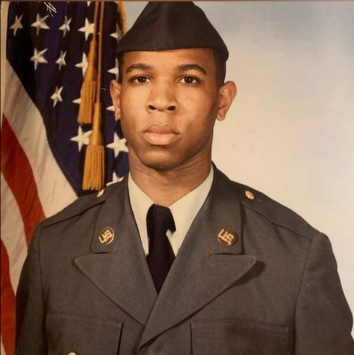 Young man in military uniform with a cap, standing in front of an American flag, looking serious.
