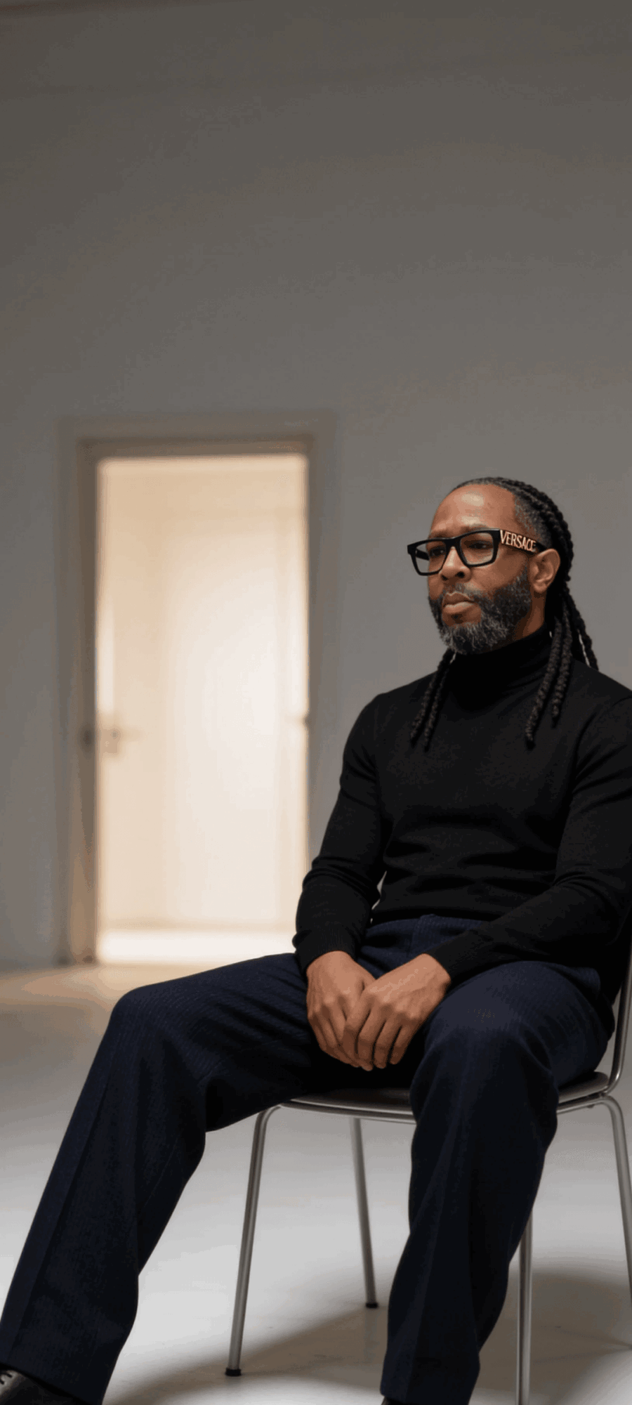 Man with glasses and dreadlocks sitting on a chair in a room with a door in the background.