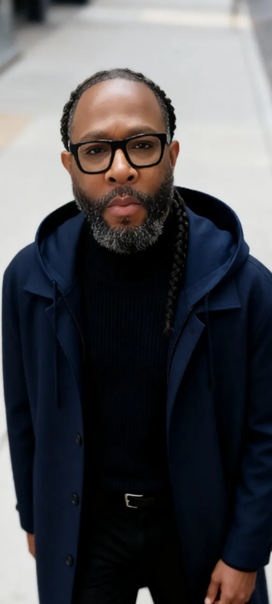 Man with glasses and beard wearing a dark jacket and hoodie, standing outdoors on a city street