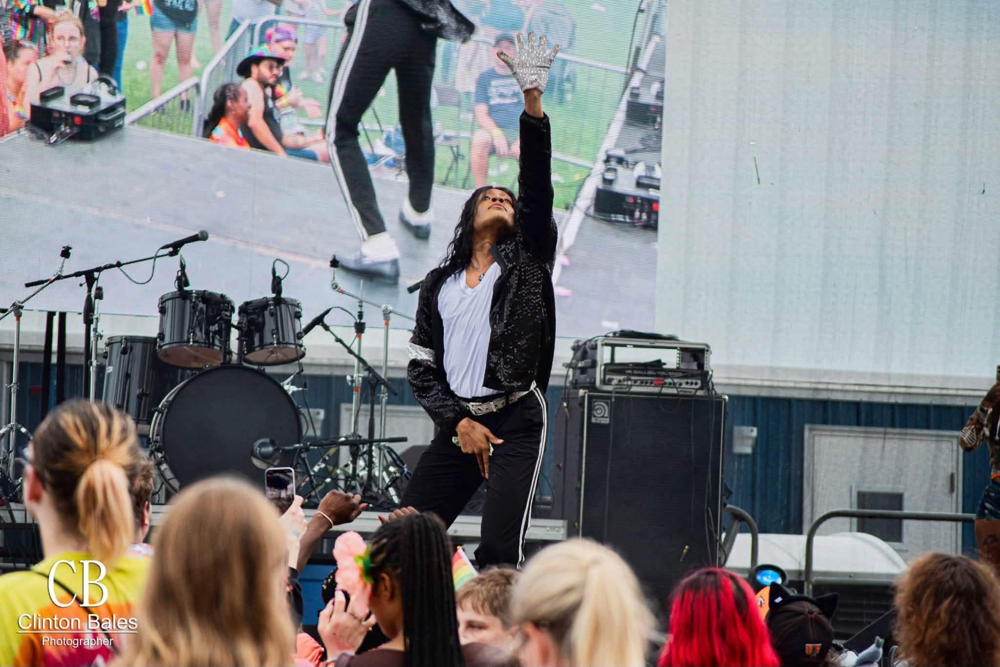 Performer on stage with microphone, raising hand, audience watching, outdoor event, musical instruments in background.