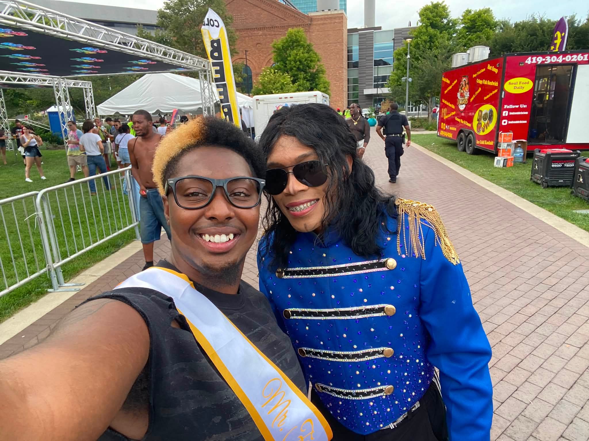 Two women smiling at an outdoor event, one wearing sunglasses and a blue jacket with gold accents, taking a selfie.
