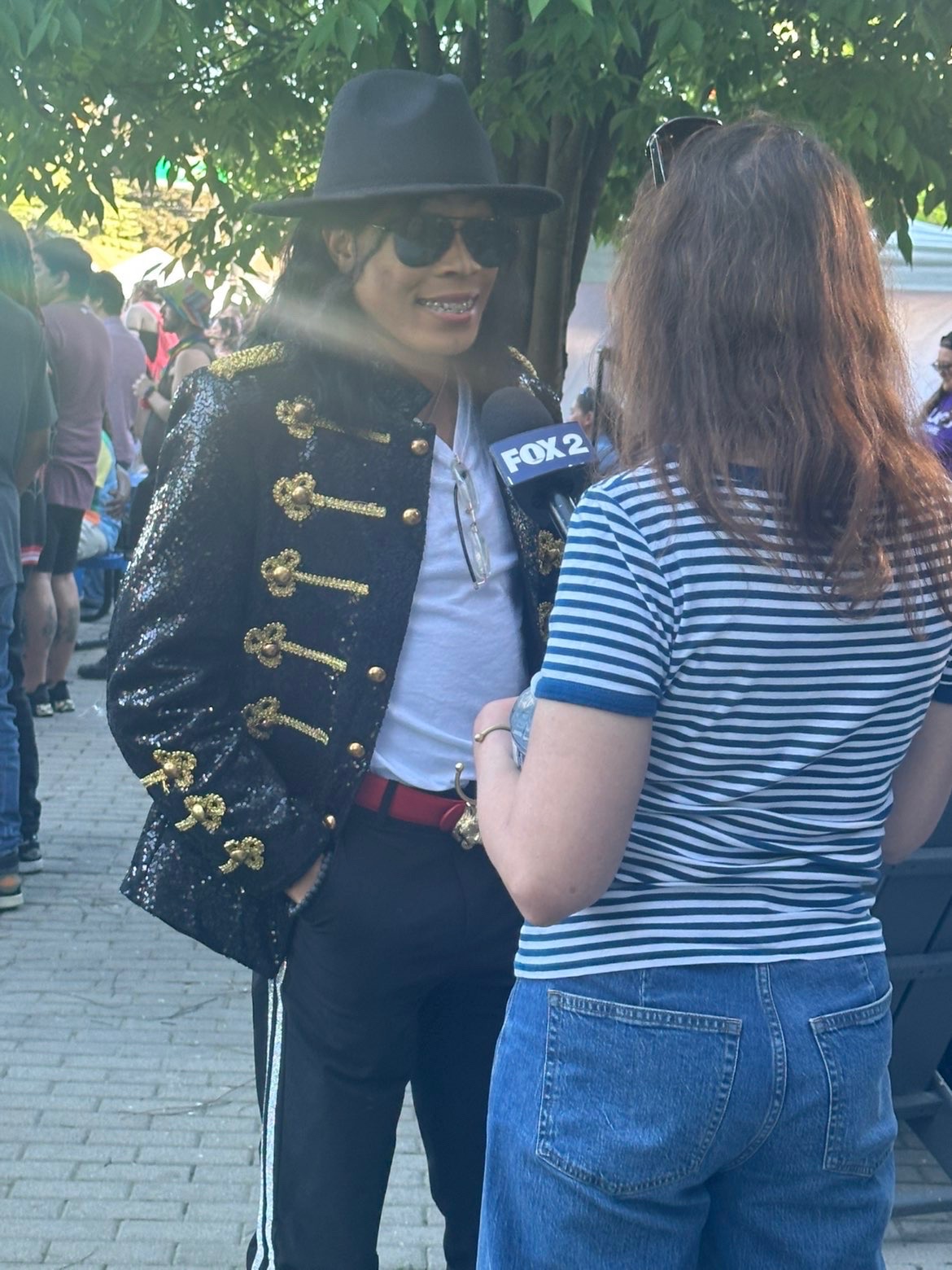 Woman wearing sunglasses, a black hat, and a black jacket with gold details, being interviewed outdoors by a person in a striped shirt.