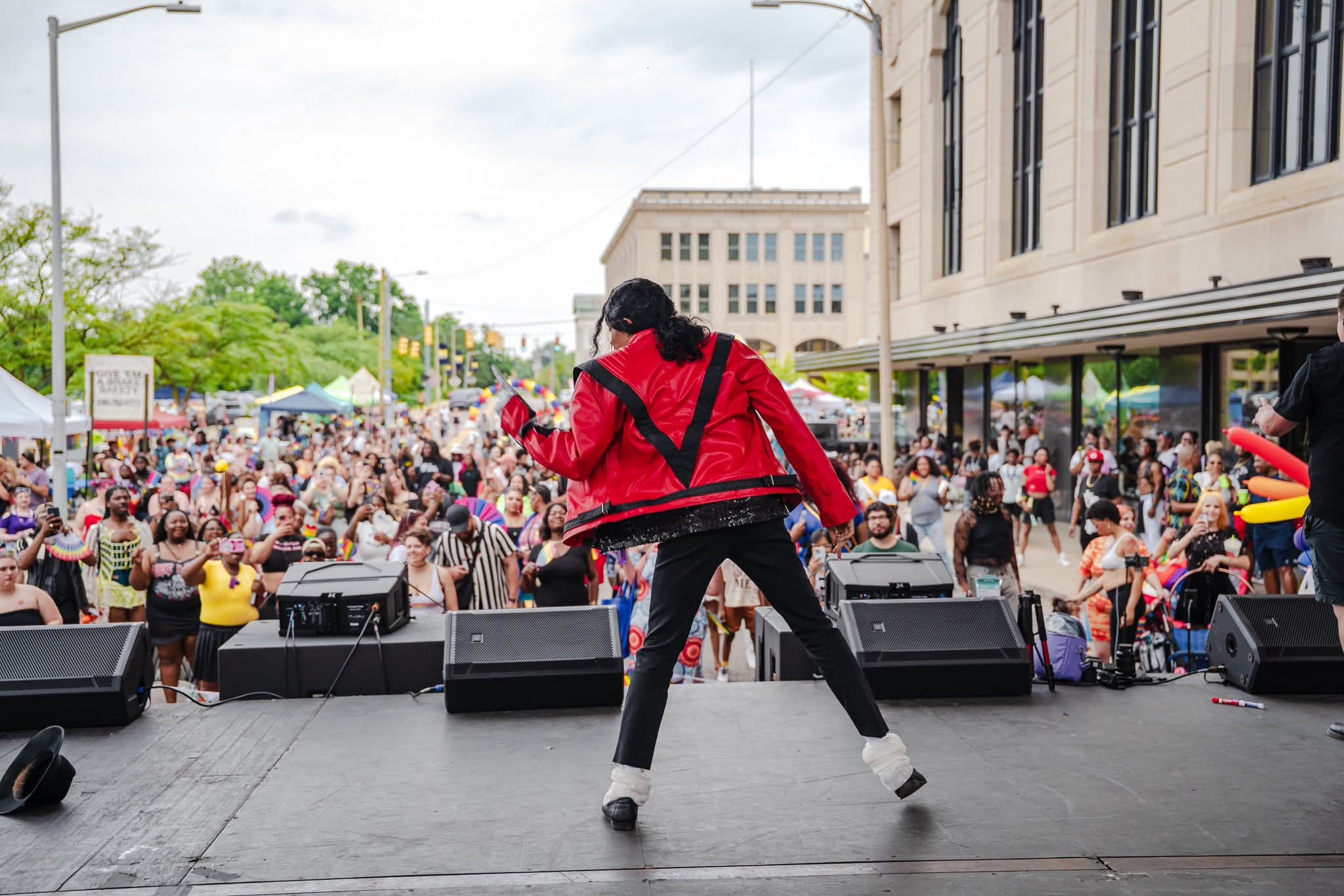 Performer on stage facing a large crowd outdoors, with buildings and trees in the background.