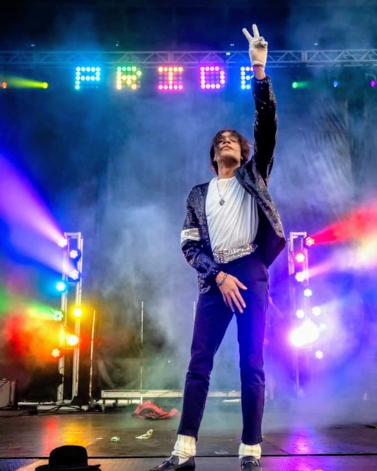 Person on stage making a peace sign with one hand, colorful lights and a sign spelling 'PRIDE' above.