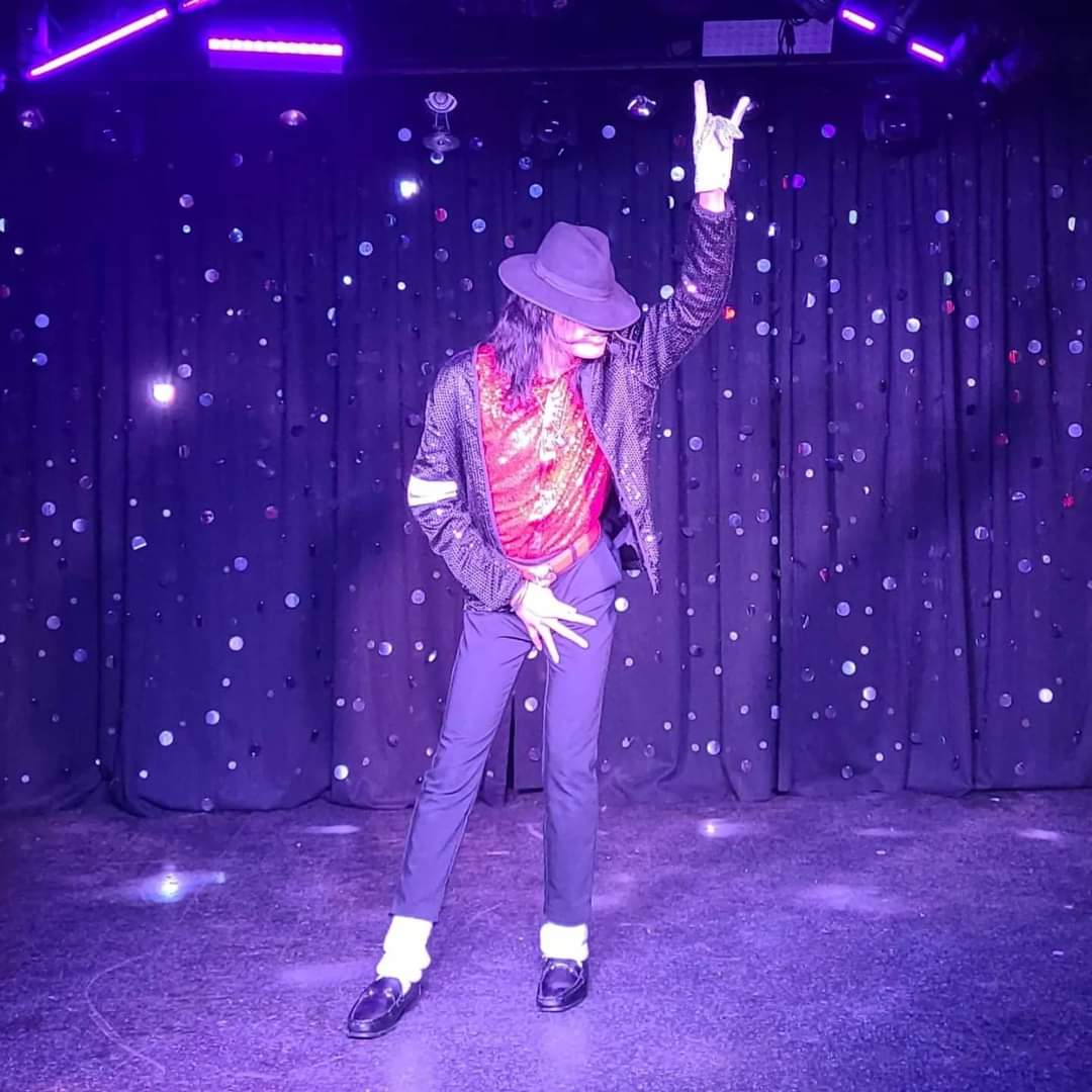 Person performing on stage with a curtain background, wearing a hat, colorful shirt, and dark pants, raising one hand.