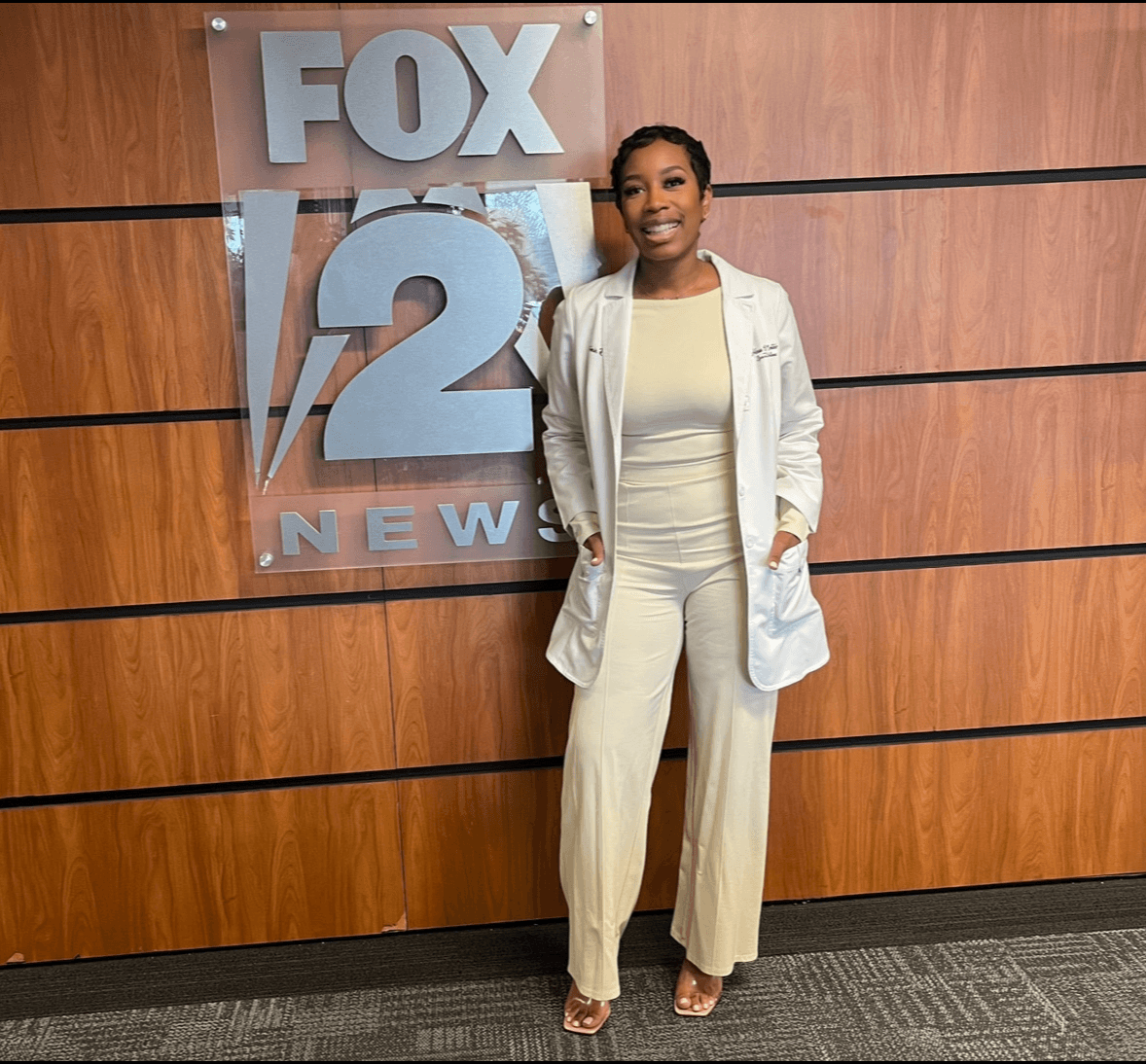 Woman in cream-colored outfit standing in front of FOX 2 News sign on wooden wall, smiling, with hands in pockets.