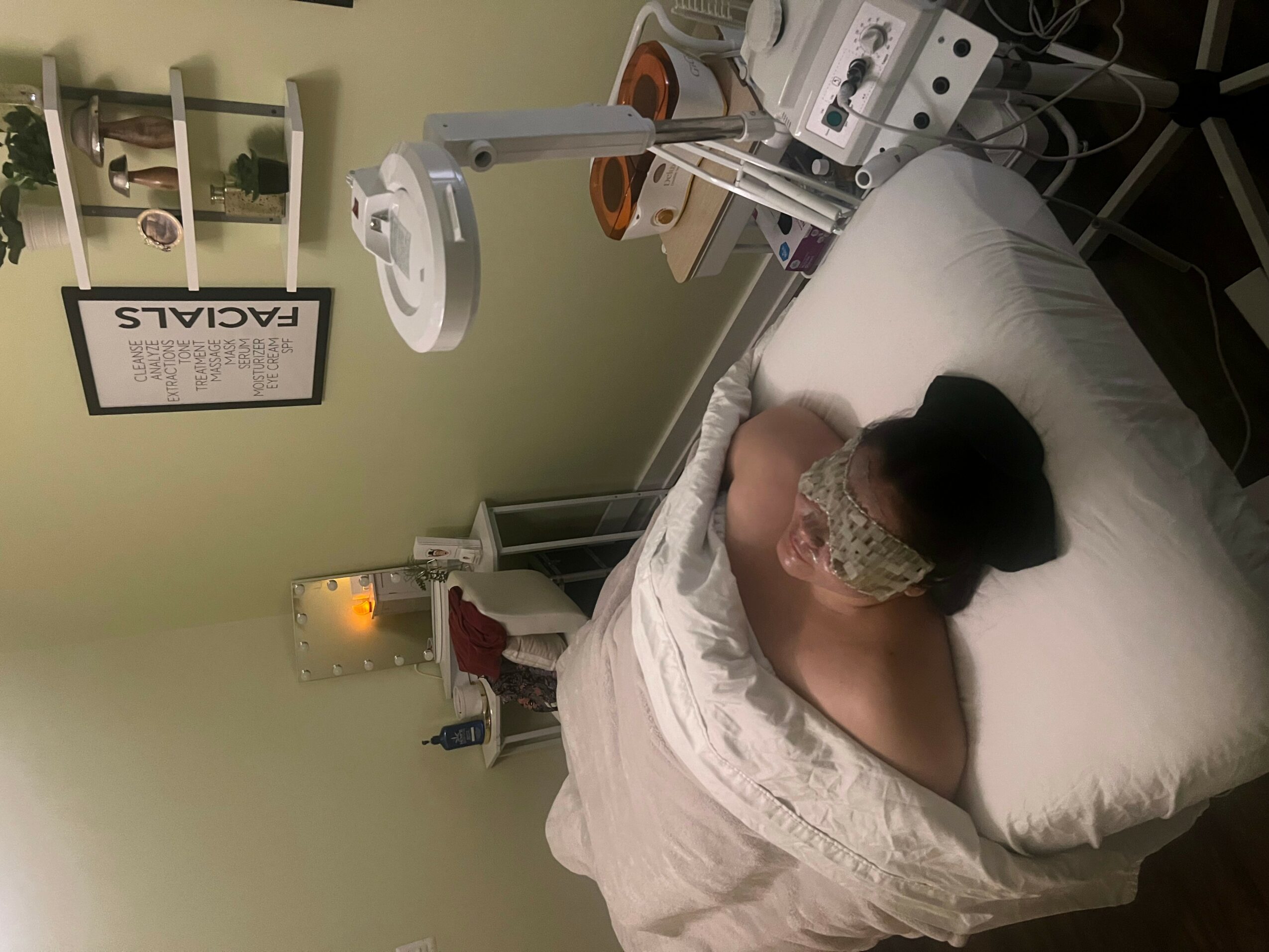 Person lying on hospital bed with medical equipment nearby, facing away from the camera, in a clinical room.