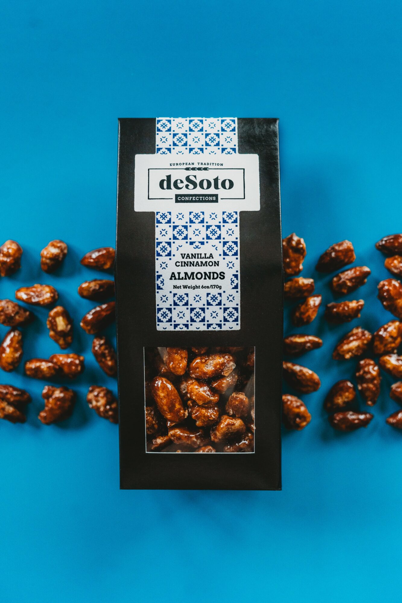 Chocolate-covered almonds in a black box with a blue and white patterned label, surrounded by scattered almonds on a blue background.