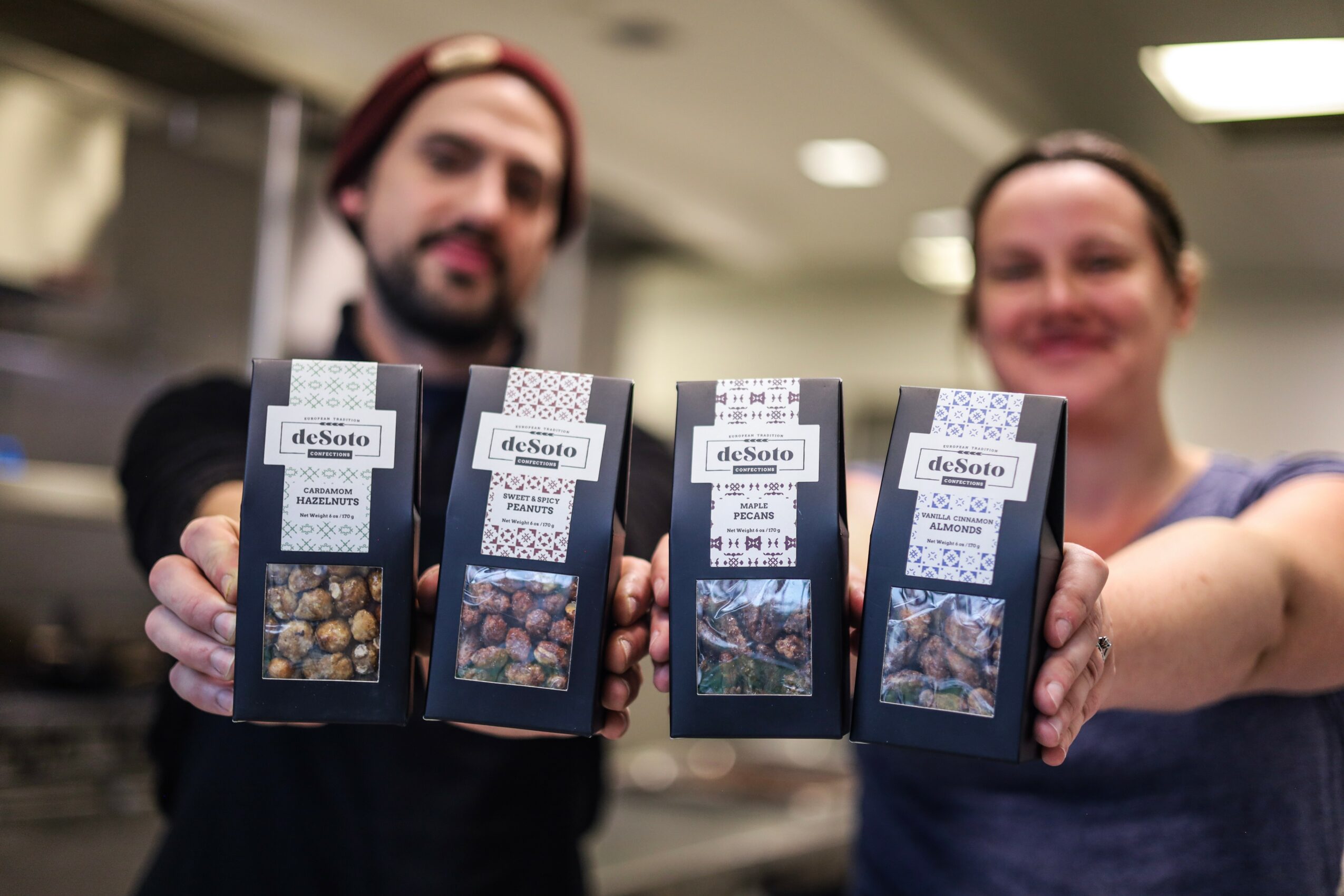 Two people hold boxes of chocolates in a store, with blurred background and smiling faces.