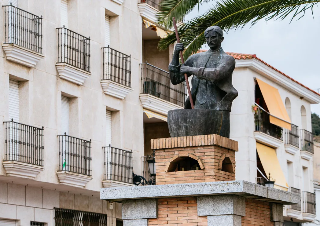 Statue of a seated woman holding a palm leaf, on a brick pedestal in front of a building with balconies.