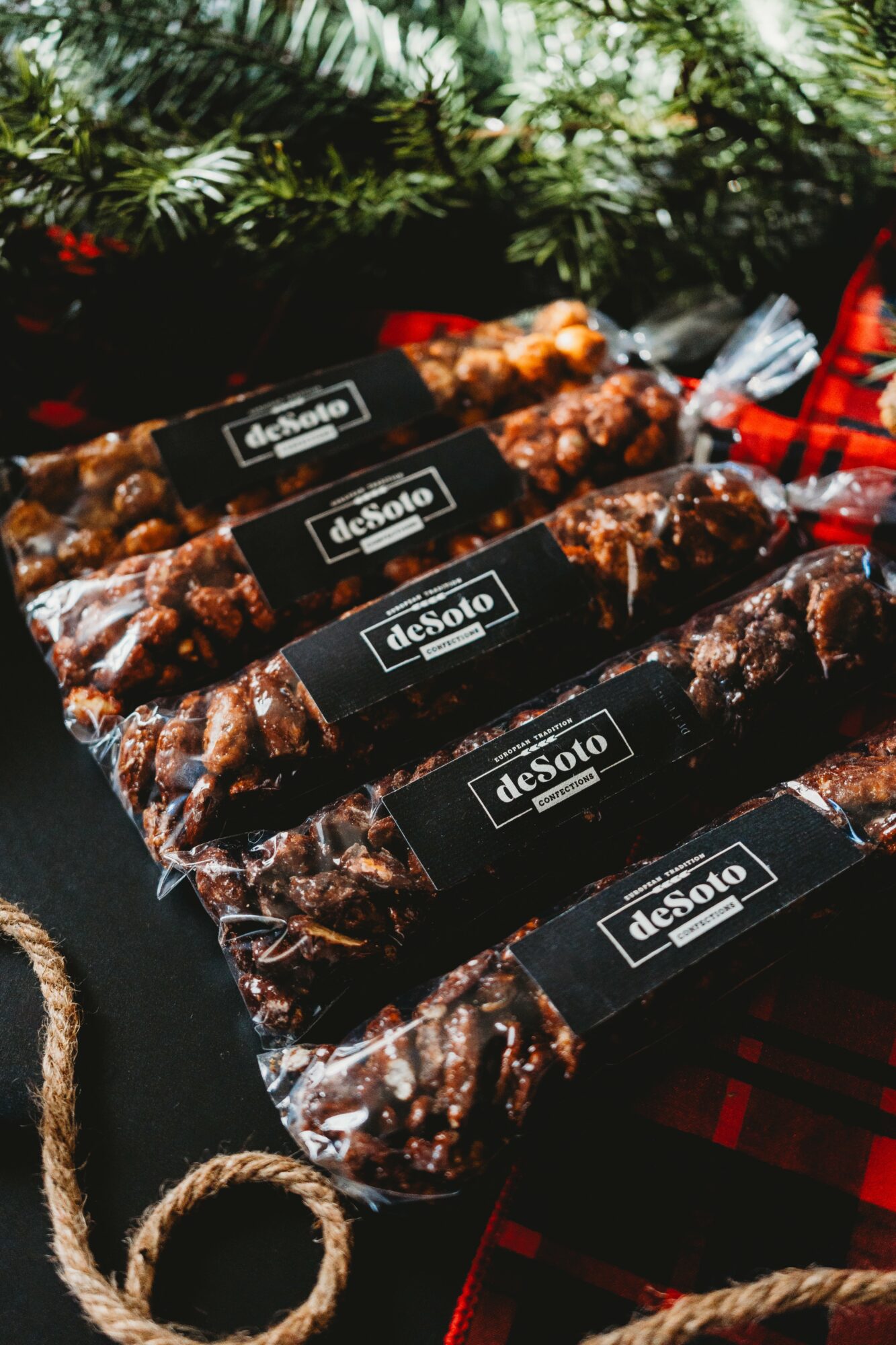 Several packages of sausage arranged on a dark surface with a red plaid cloth nearby, surrounded by green pine branches.