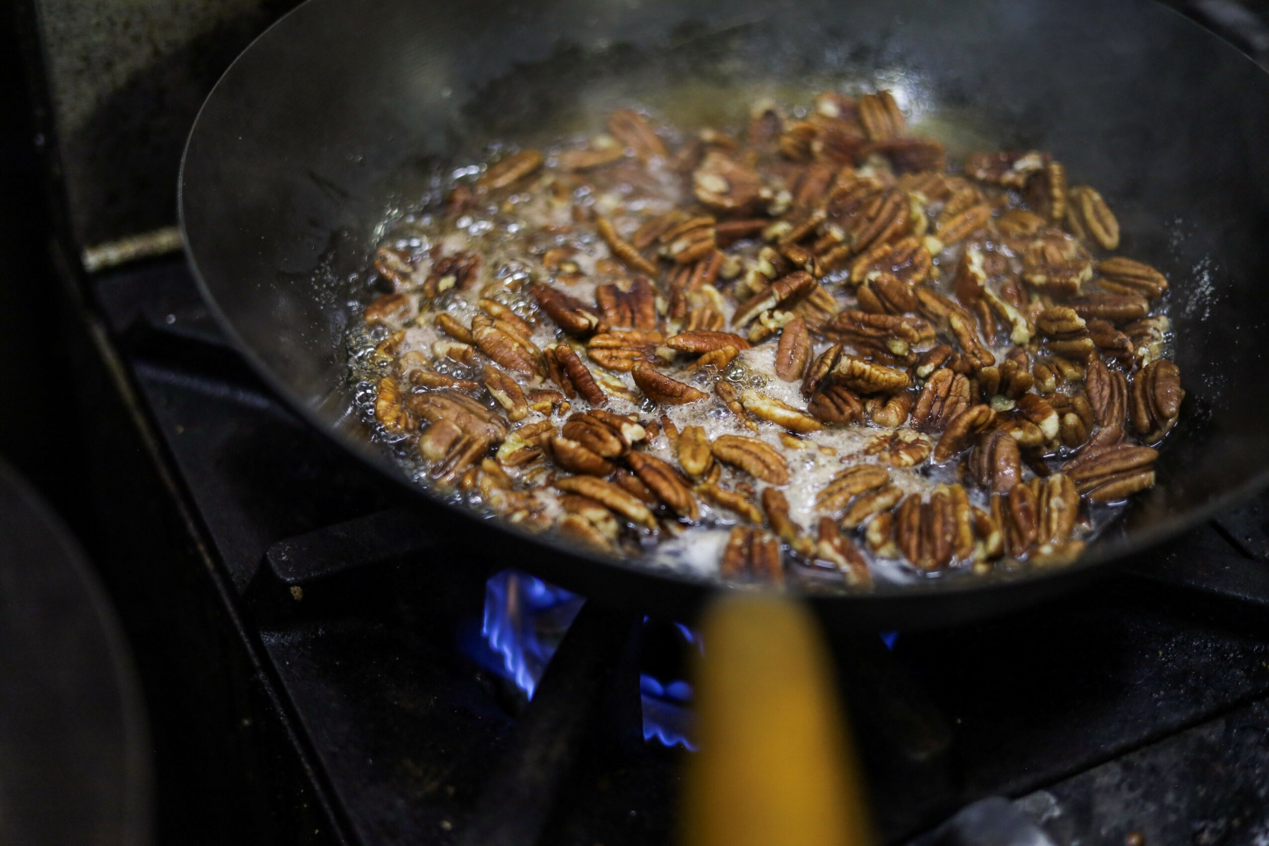 Pecans cooking in a frying pan on a stove with blue flame visible, steam rising from the pan.