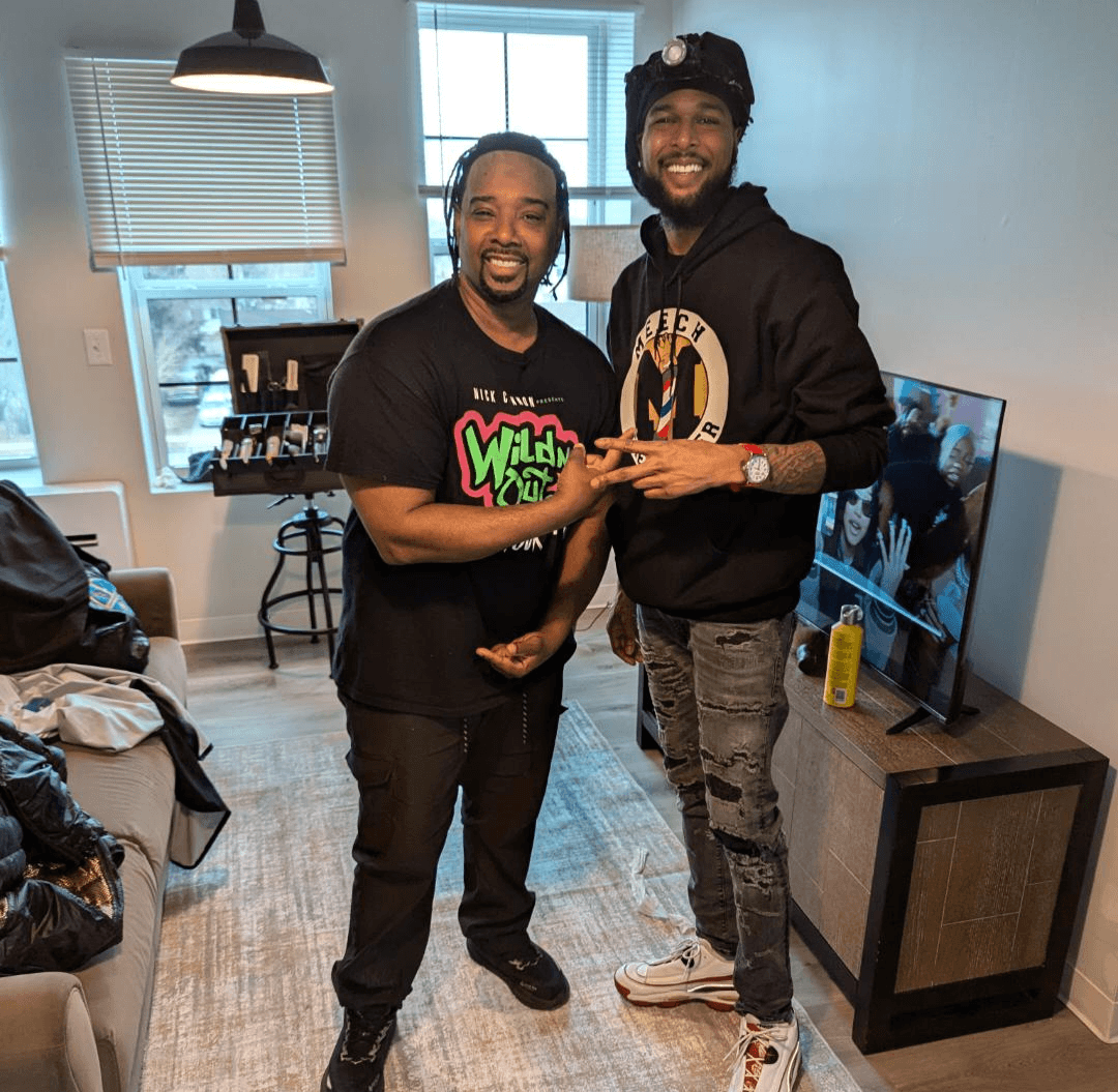 Two men smiling and shaking hands in a living room, with a TV and window in the background.