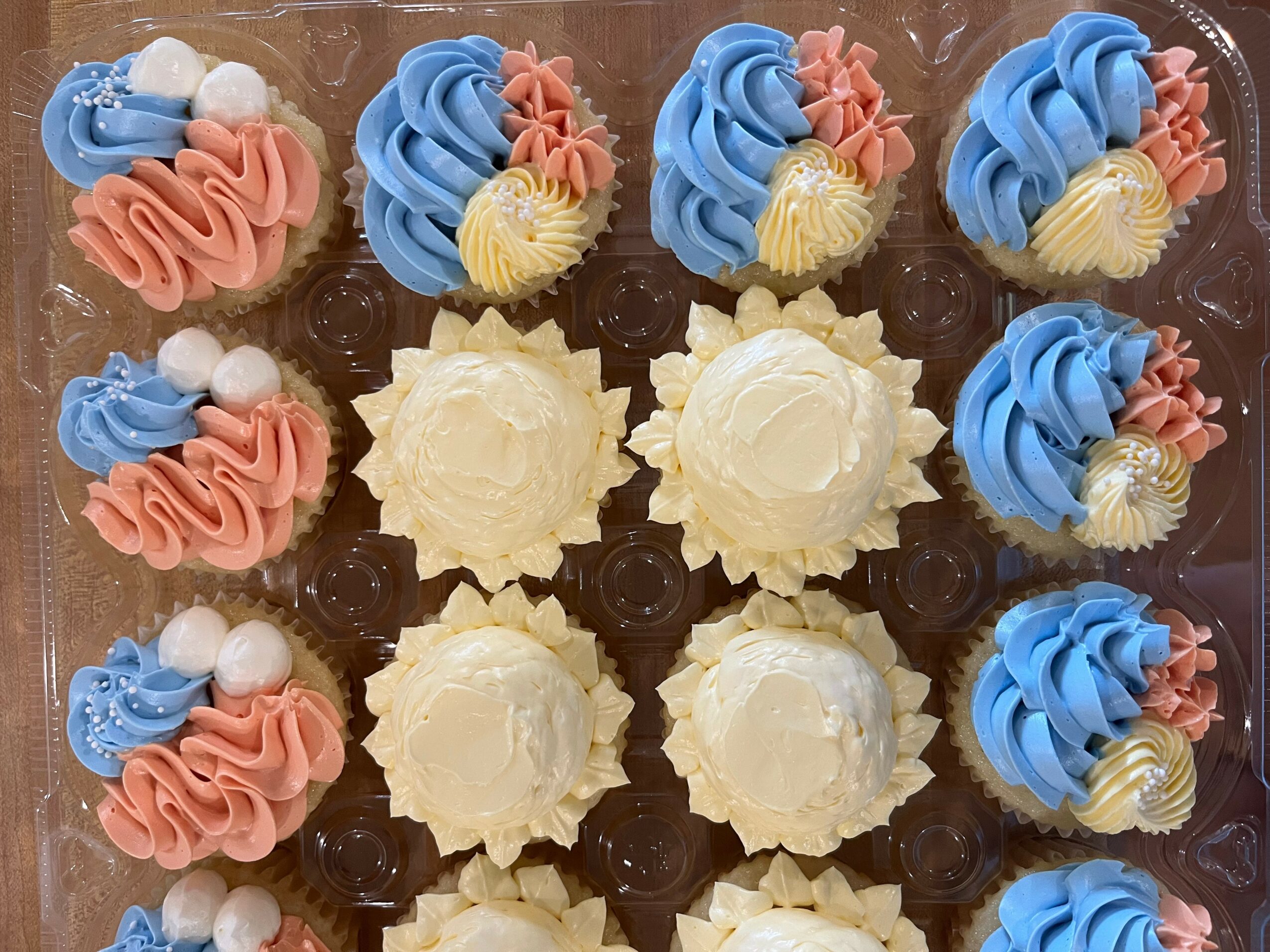 Assorted cupcakes with blue, pink, and white frosting, decorated with small flowers and pearls, arranged on a tray.