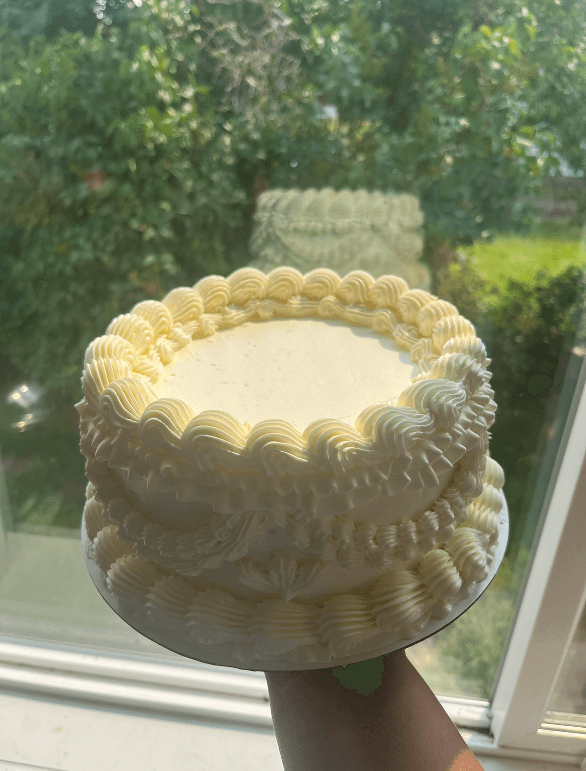 Cake with white frosting and decorative piping, viewed through a window with greenery outside