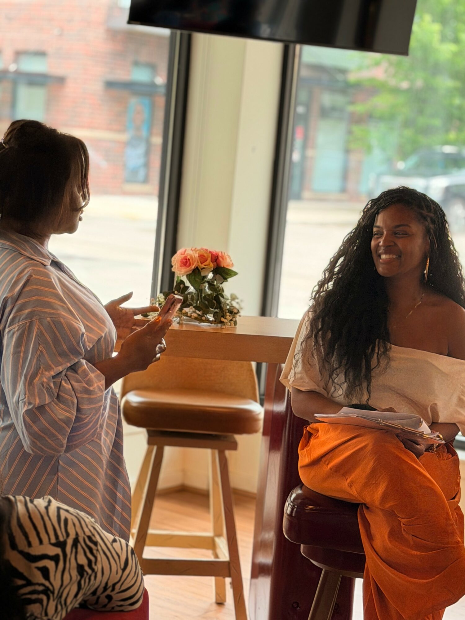 Two women sitting and talking inside a cafe with large windows, a flower arrangement, and a zebra-patterned bag, smiling.