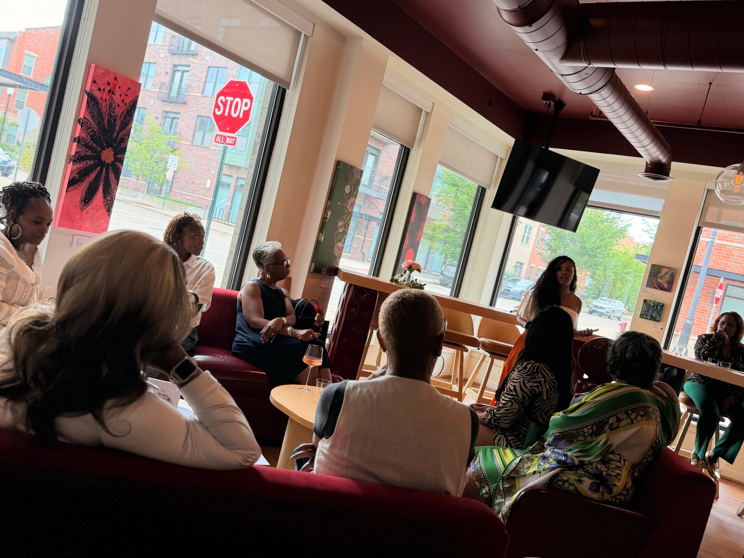 Group of people seated in a room with large windows, listening to a speaker at the front.