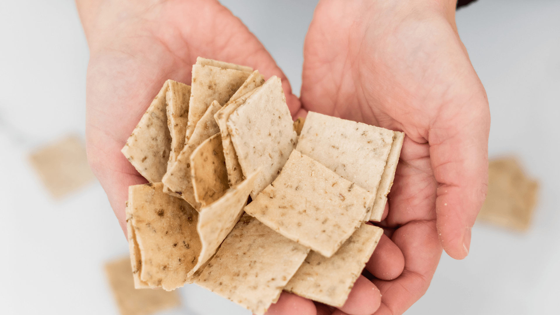 Hands holding broken pieces of flatbread or crackers, with some pieces scattered in the background.