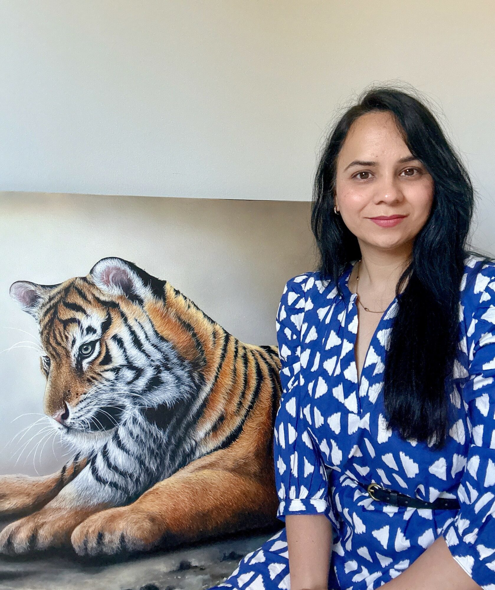 Woman with long dark hair sitting next to a tiger cub on a couch, wearing a blue and white patterned dress.
