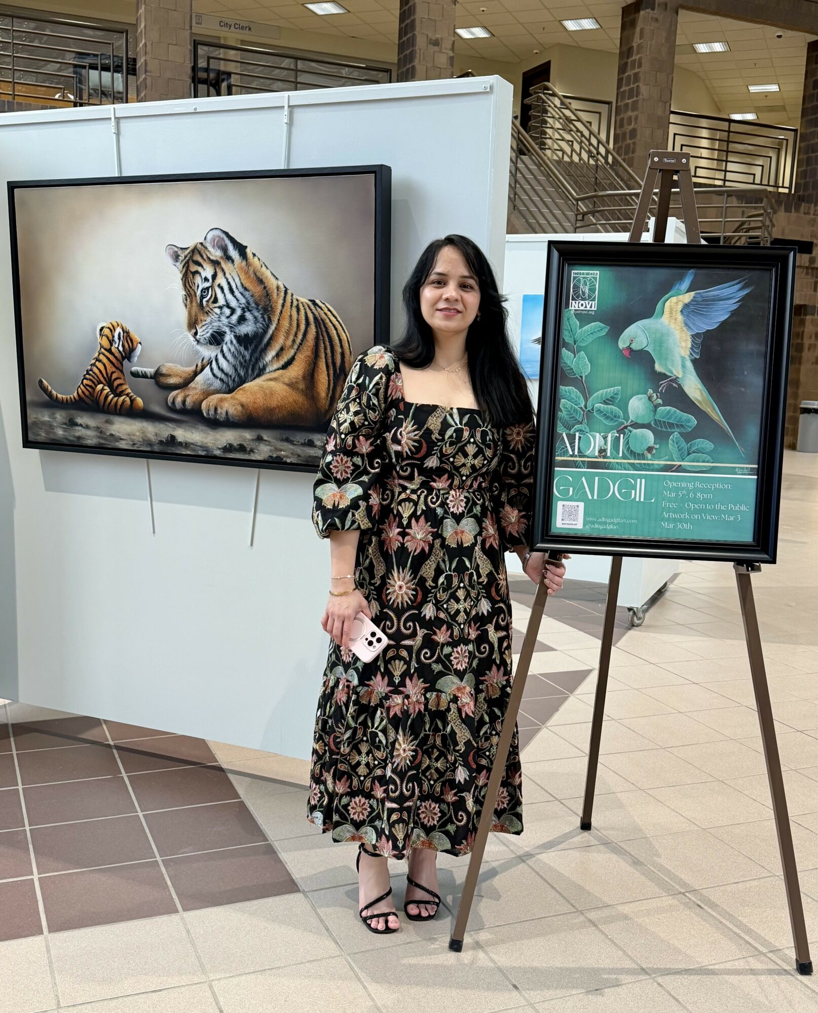 Woman standing next to two framed animal paintings, one of a tiger and a cub, and a colorful bird, in an indoor setting.