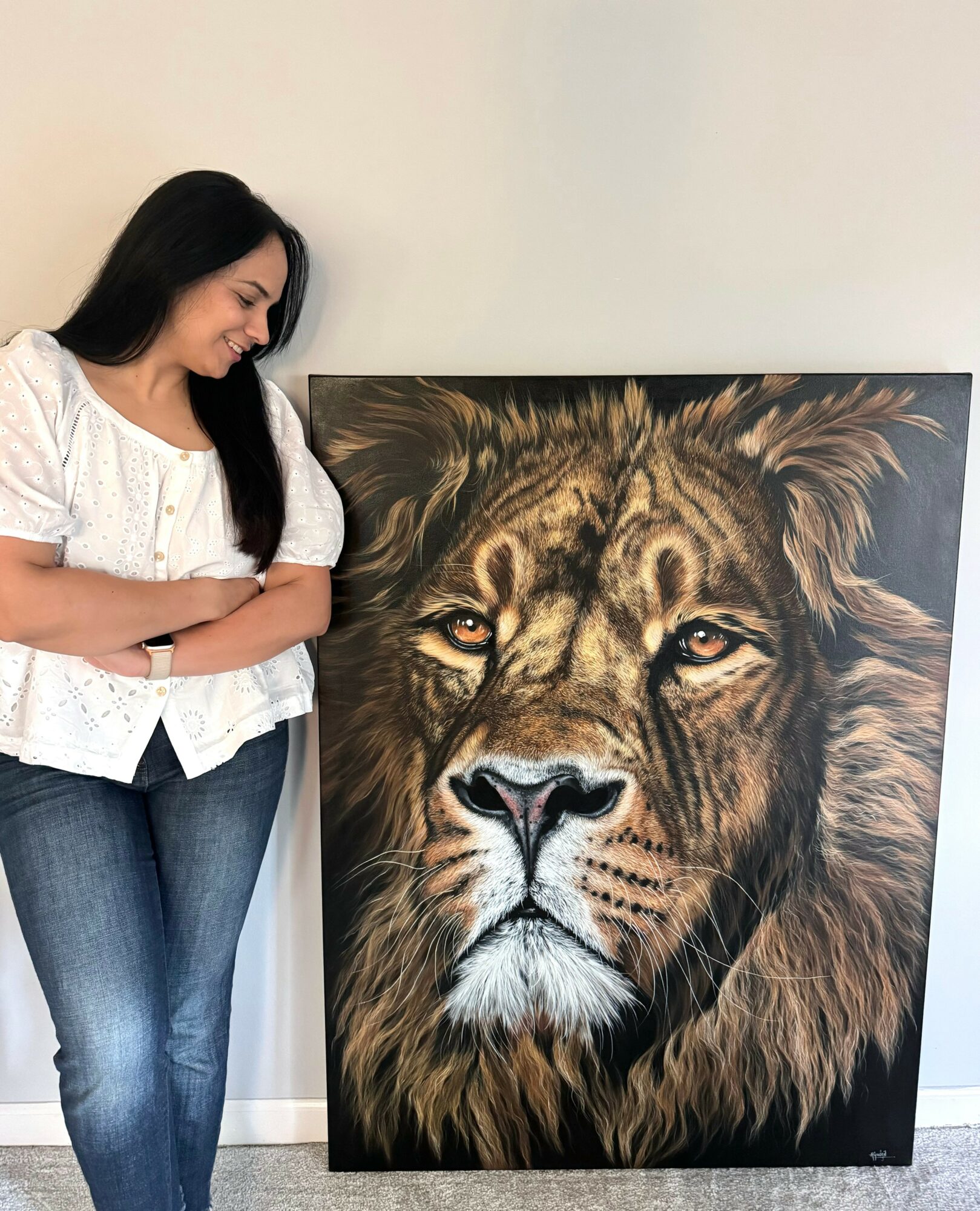 Woman with long dark hair smiling and looking at a large lion painting, standing with arms crossed, against a plain wall.