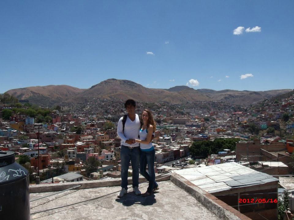 Two people stand on a rooftop overlooking a city with hills in the background under a blue sky.