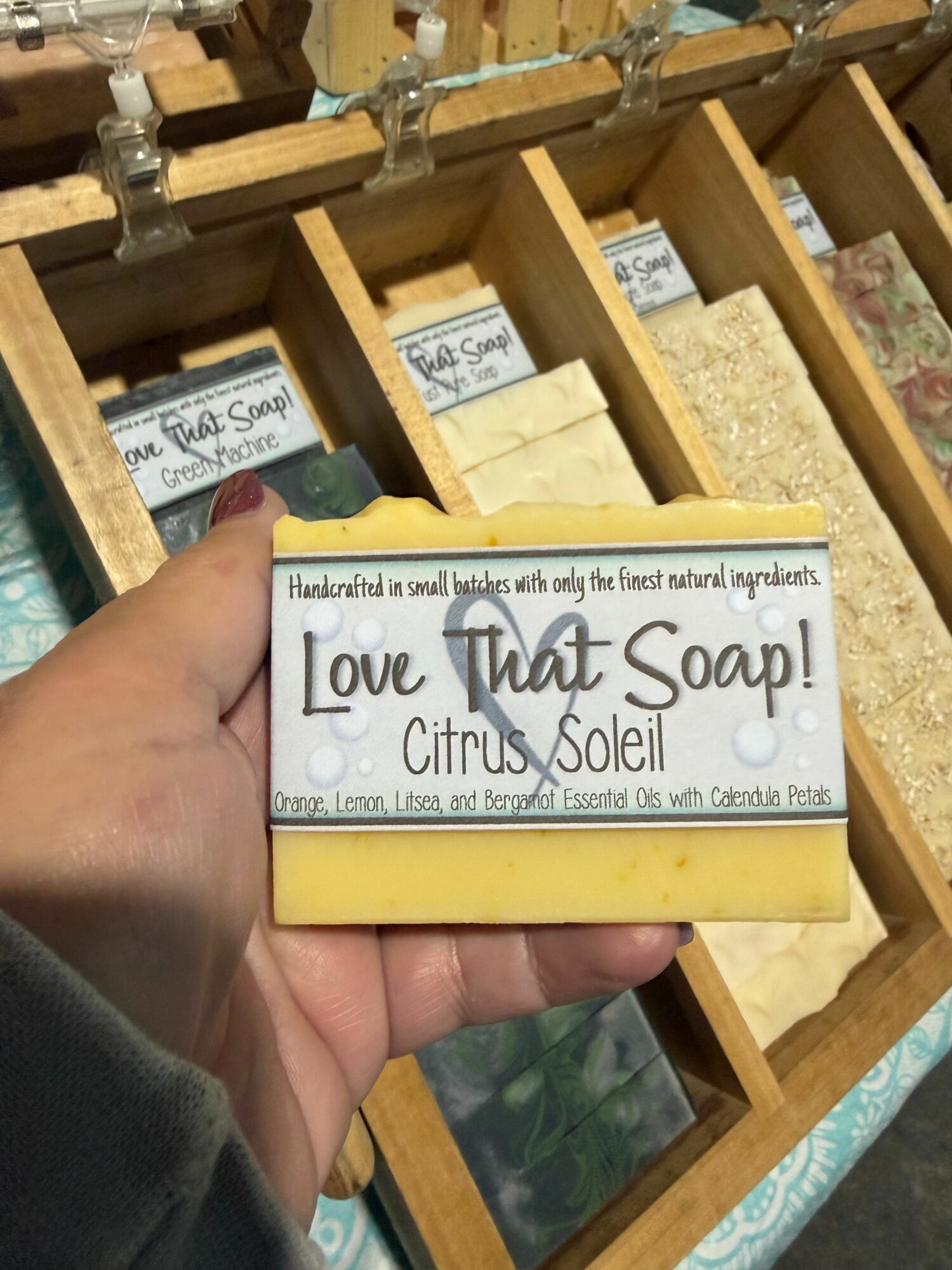 Hand holding a soap bar labeled 'Love Notes Soap' with a citrus scent, in front of a wooden display with other soap bars.