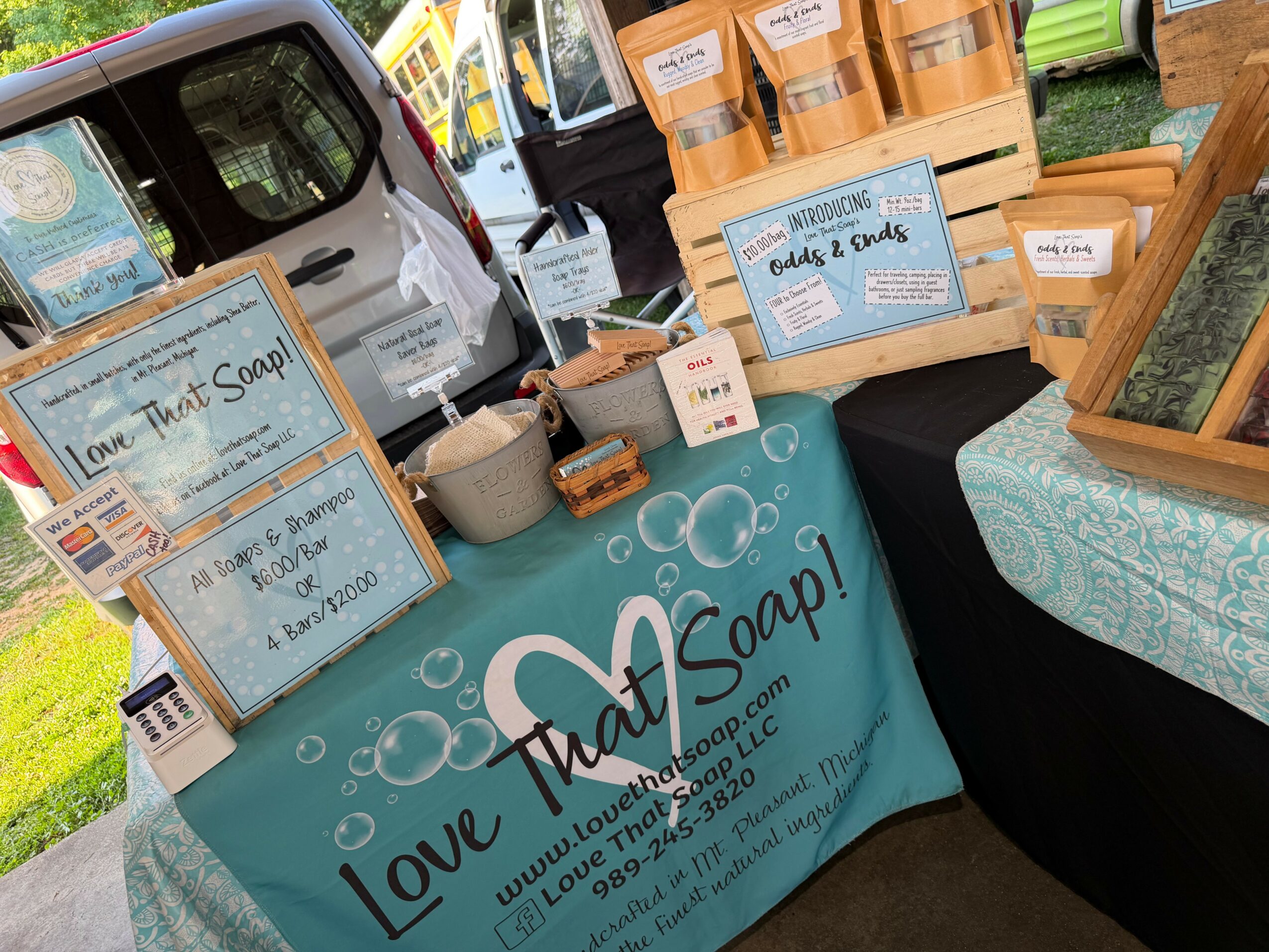 Table with soap products and signs, outdoor market setting, vehicle and grass in background.