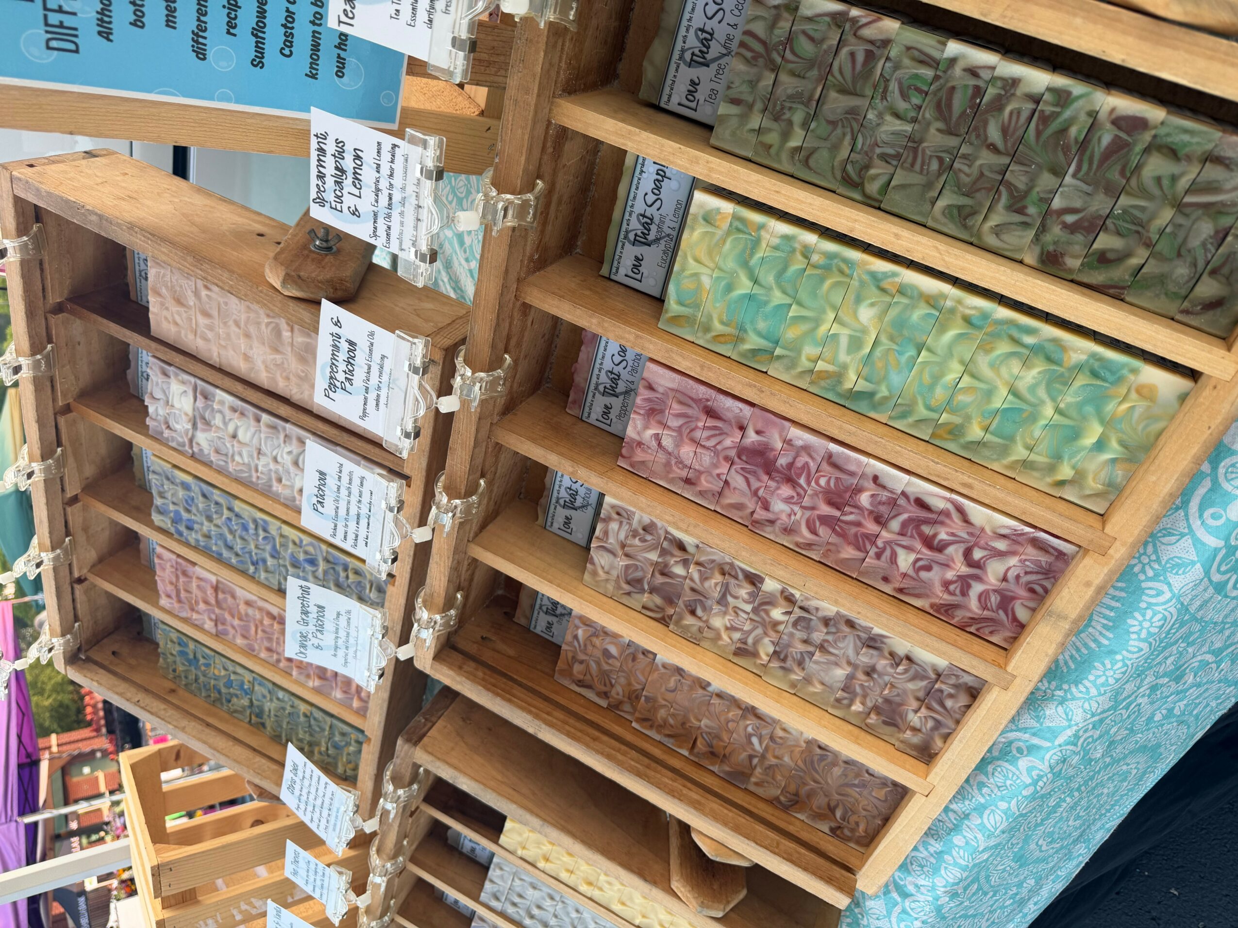 Wooden display with colorful soap bars arranged on shelves, some with labels, next to a blue patterned cloth.